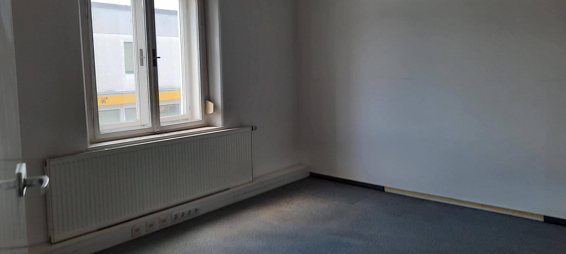 Heller Büroraum mit großem Fenster, Heizkörper und grauem Teppichboden, ideal für Arbeitsplätze.