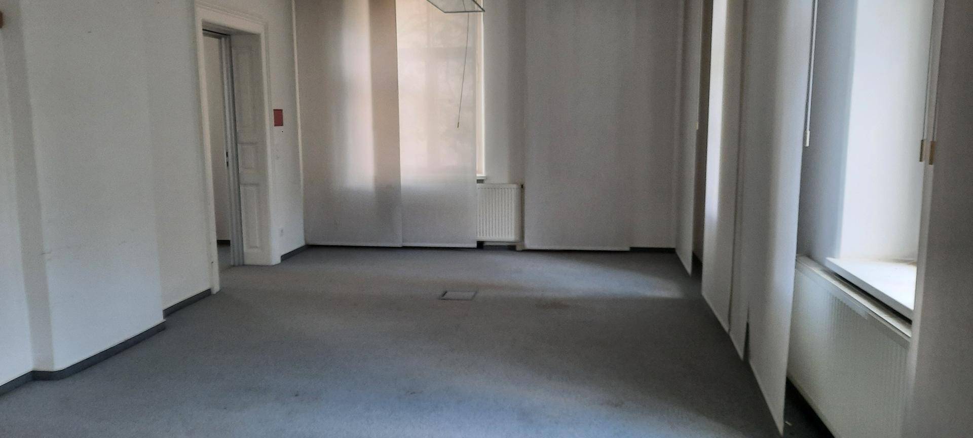Geräumiger, leerer Büroraum mit mehreren Fenstern und grauem Teppichboden, bietet viel Tageslicht.