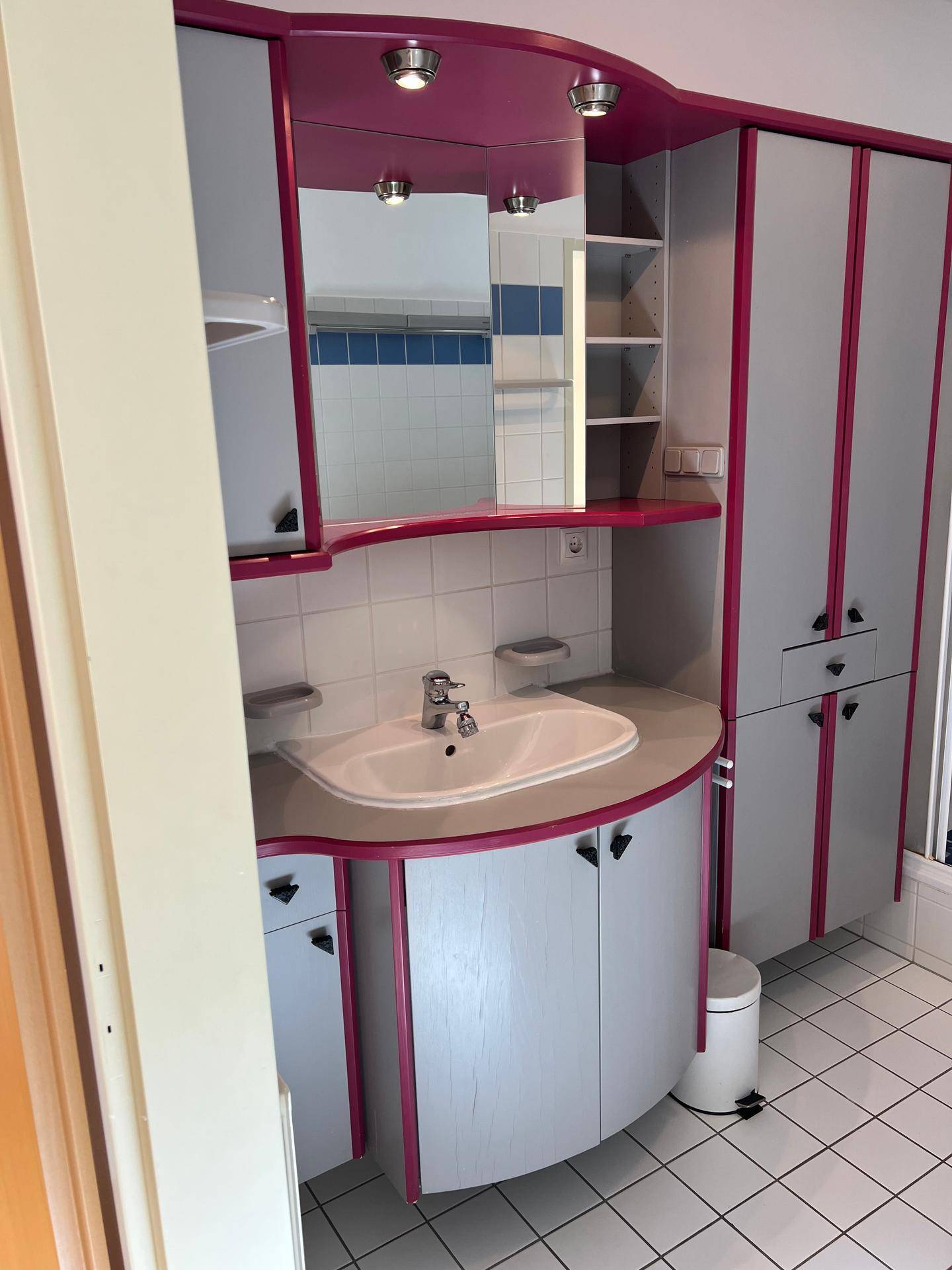 Badezimmer mit Waschtisch, Spiegel und farbigen Einbauschränken in Grau und Pink.