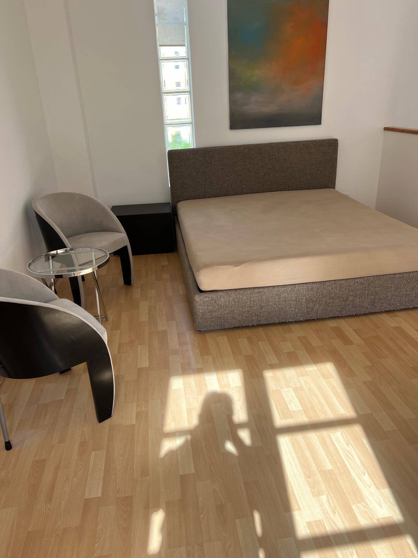 Schlafzimmer mit Bett, zwei Stühlen und Laminatboden, hell durch ein Fenster mit Glasbausteinen.