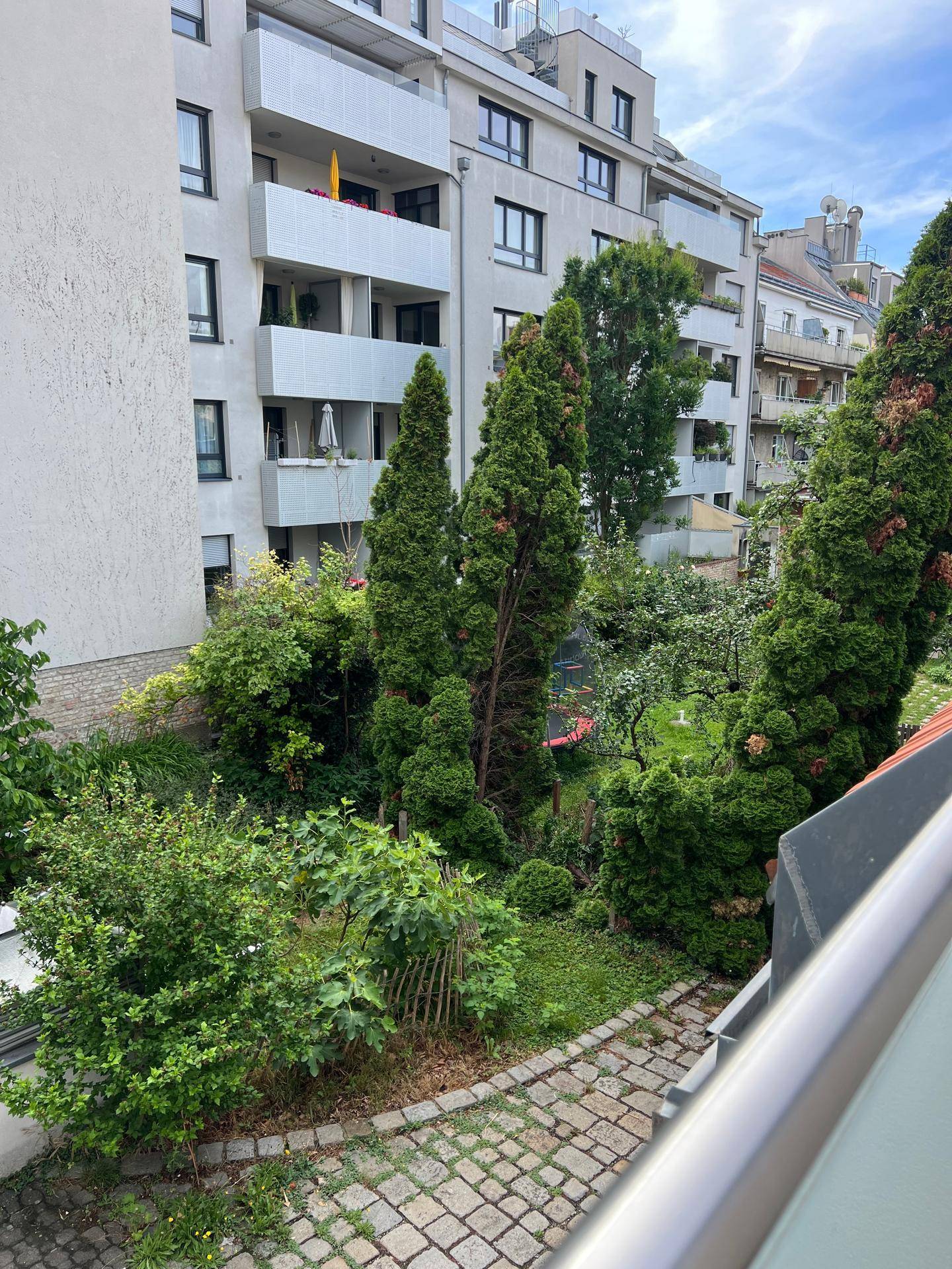 Blick auf einen gepflegten Gemeinschaftsgarten mit Bäumen, Sträuchern und einem gepflasterten Weg.