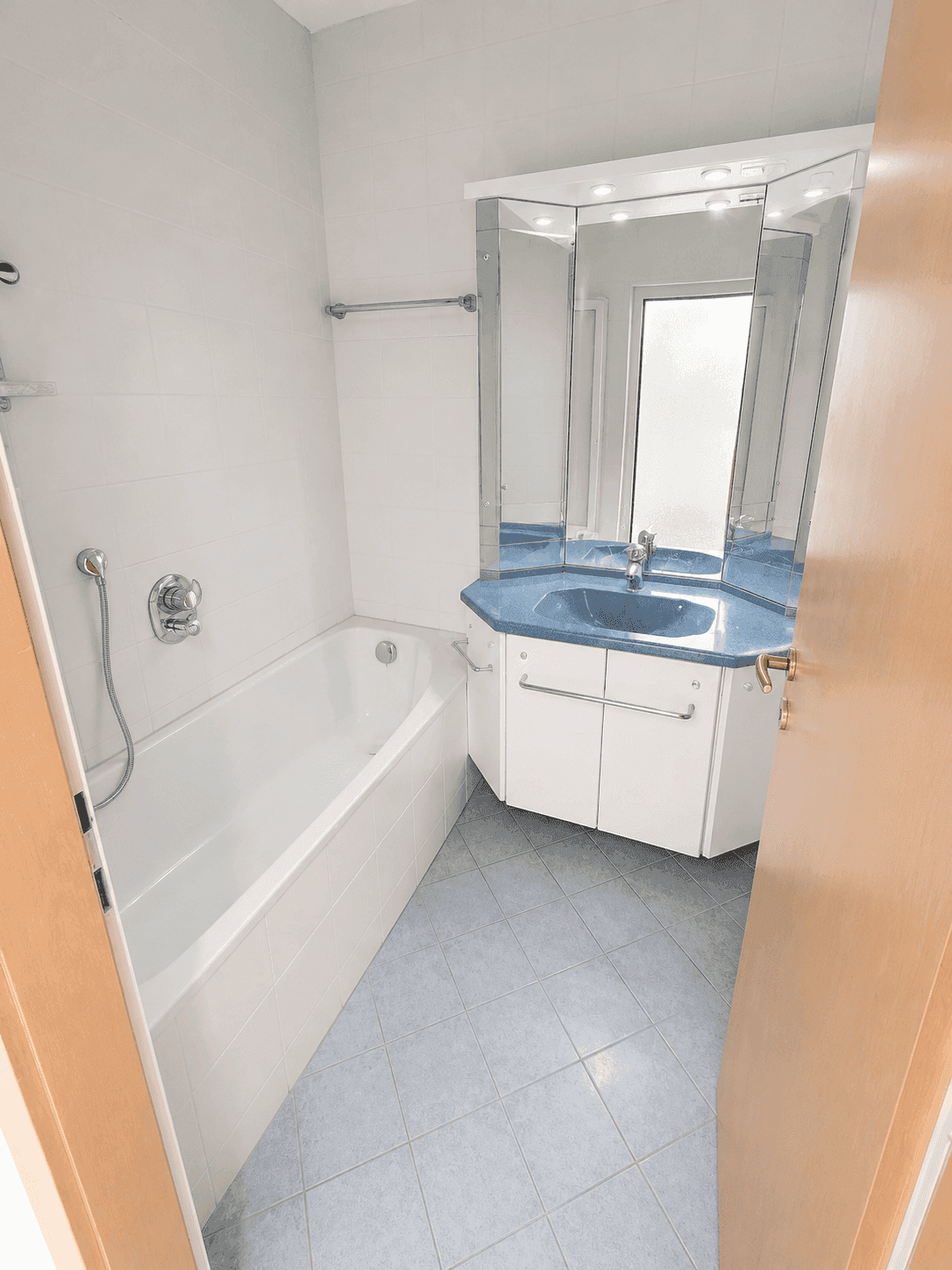 Badezimmer mit Badewanne, blauem Waschbecken und Spiegelschrank, gefliest in Weiß und Hellblau.