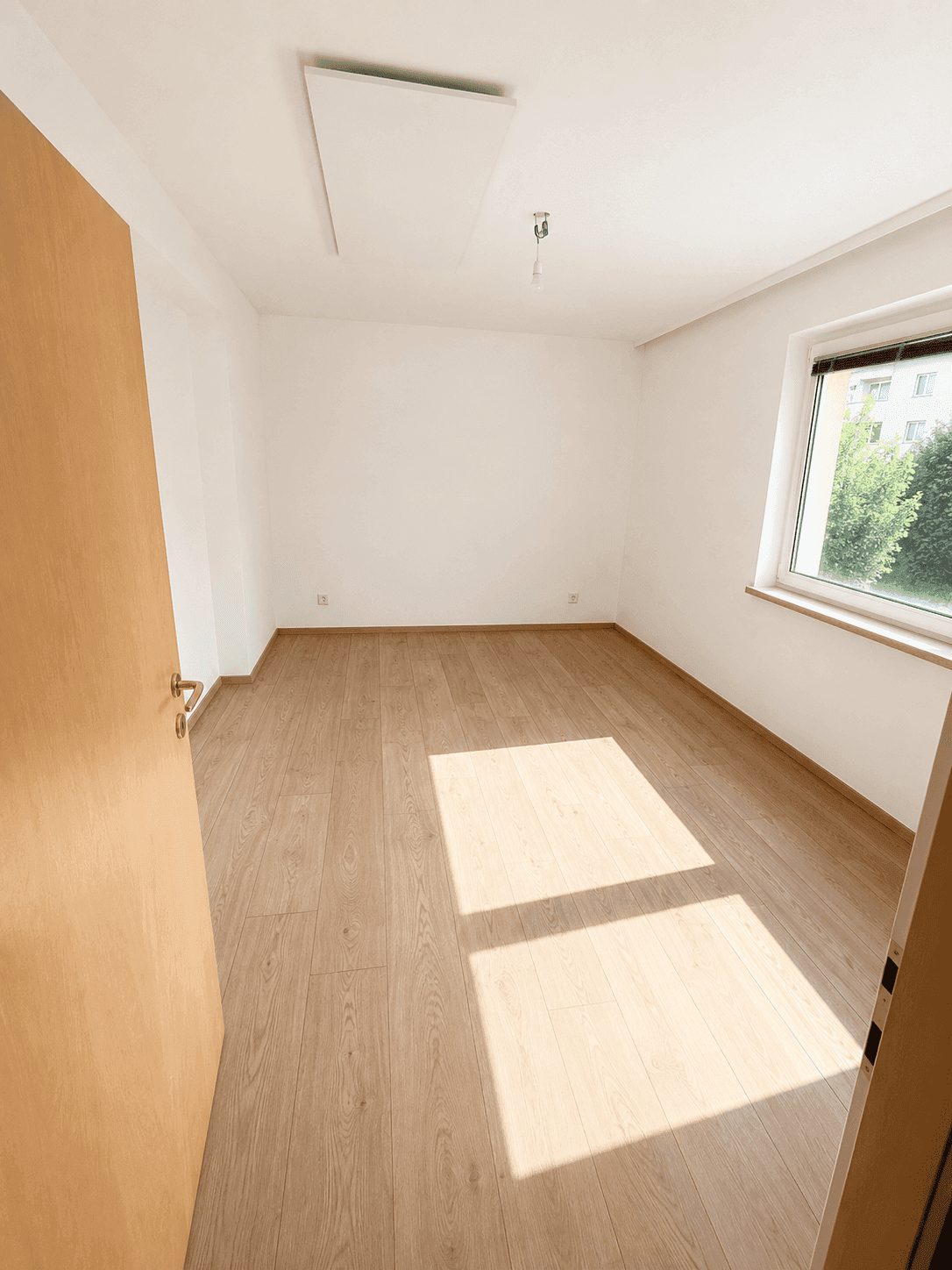 Sonniger Raum mit hellem Holzplankenboden und einem Fenster, das viel Tageslicht hereinlässt.