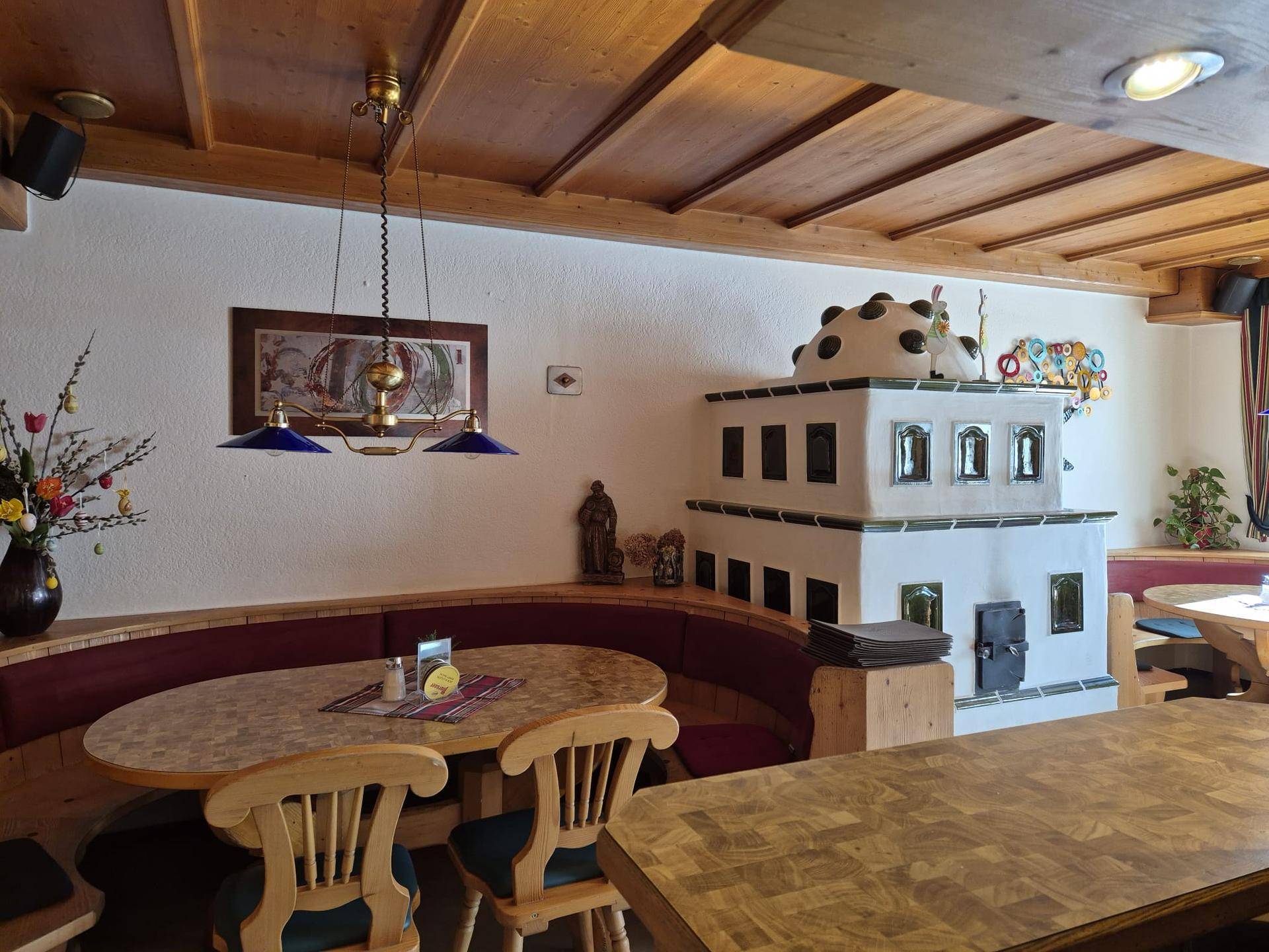 Rustikaler Innenraum eines Restaurants mit Holzbalkendecke und traditionellem Kachelofen als Blickfang.