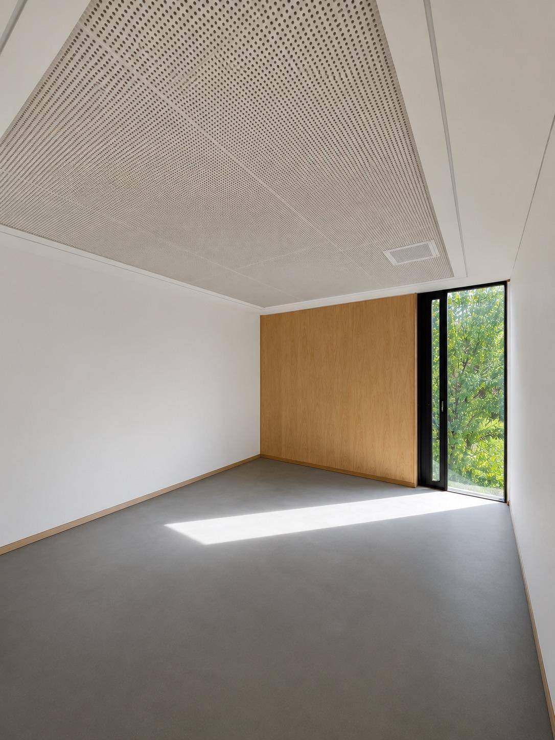 Heller, leerer Raum mit Holzakzentwand, Betonboden, hohem Fenster und perforierter Decke.