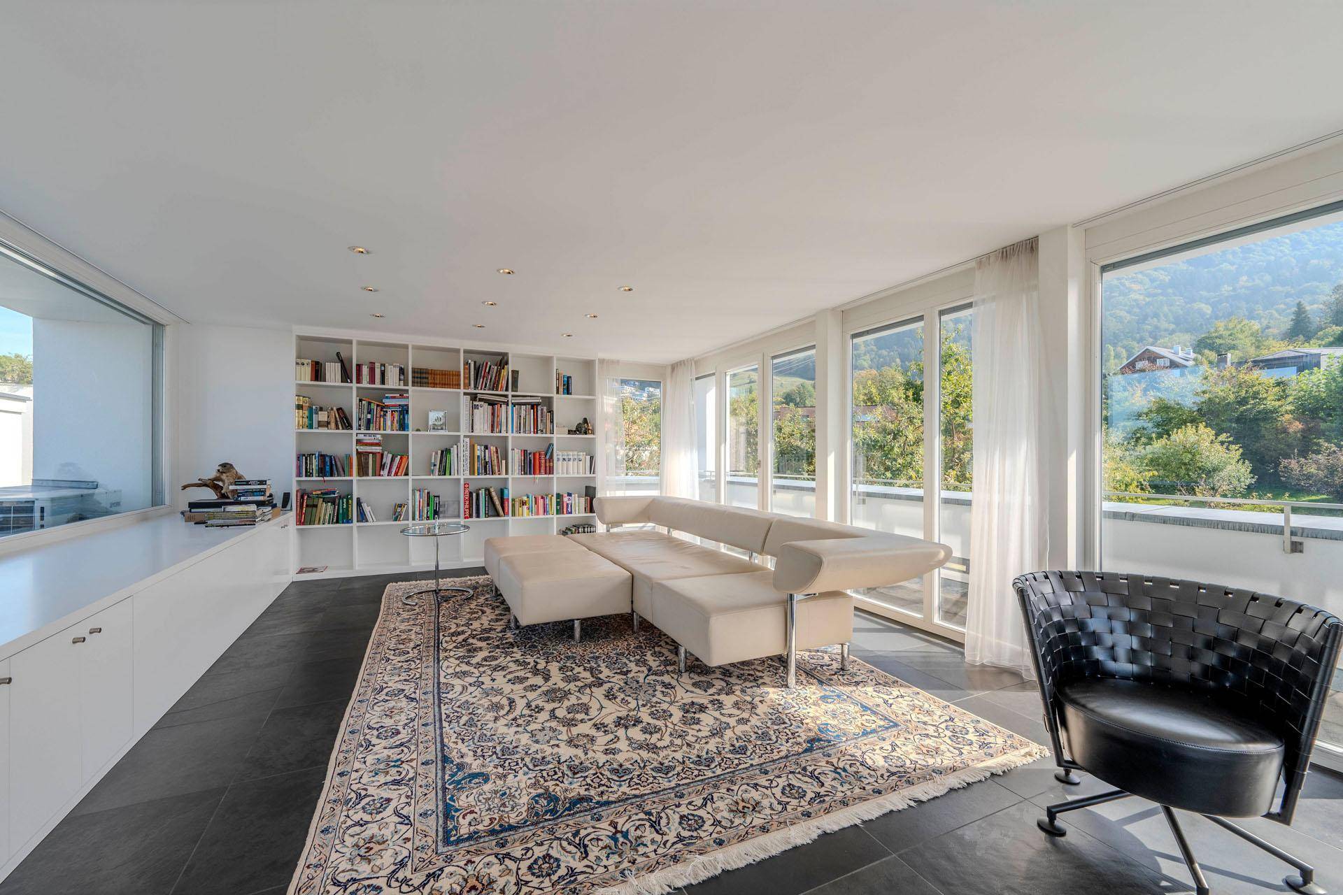 Helles Wohnzimmer mit großen Fenstern, Bücherregal und Blick auf die grüne Umgebung.