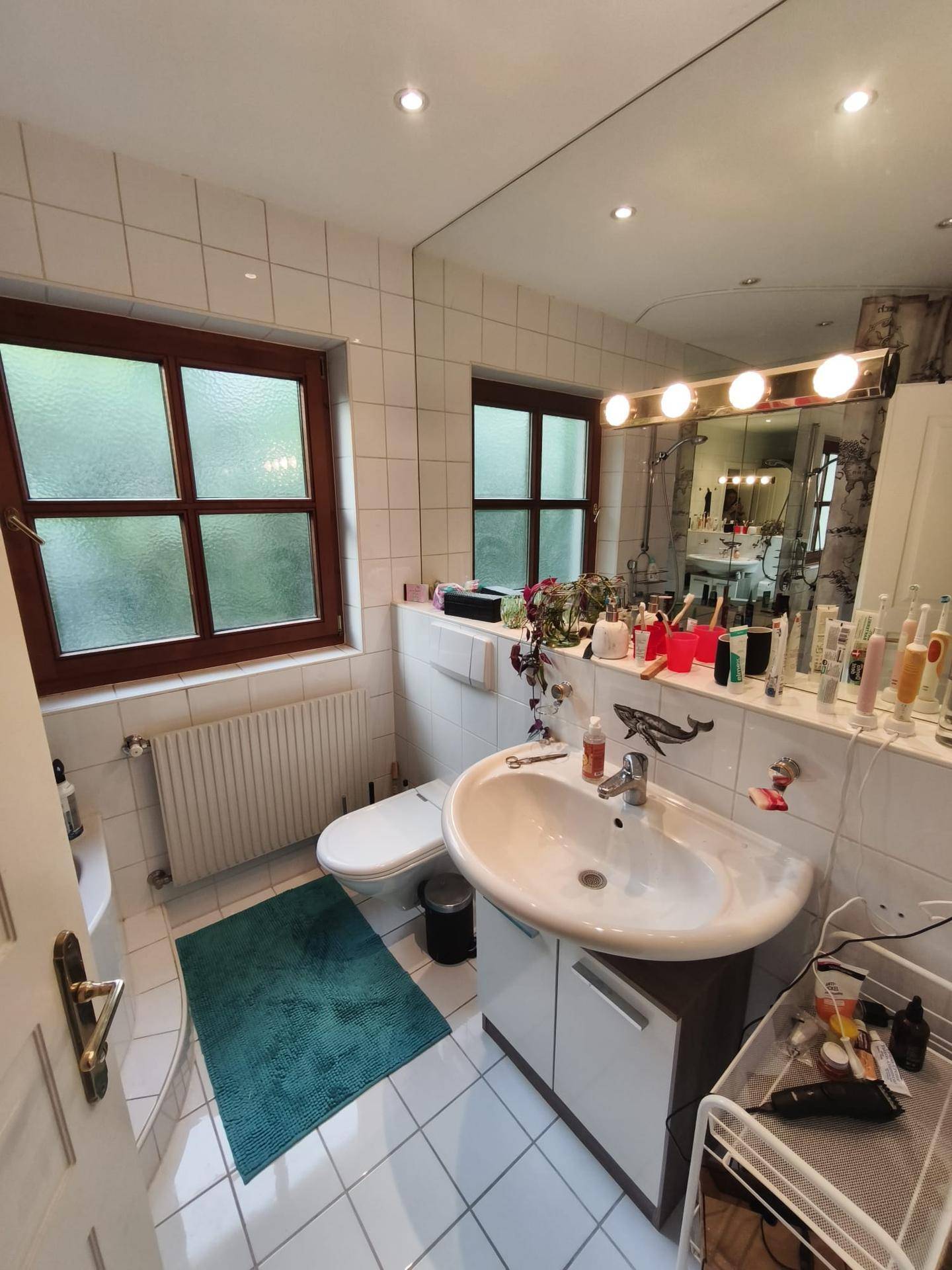 Helles Badezimmer mit weißen Fliesen, Waschbecken, Toilette und zwei Fenstern für Tageslicht.