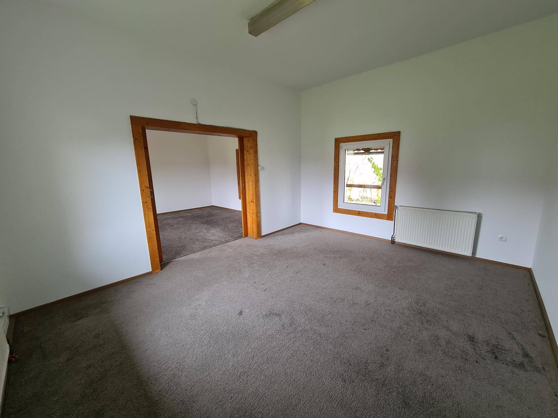 Helles Zimmer mit grauem Teppichboden, Holzfenster und offenem Zugang zu einem angrenzenden Raum.