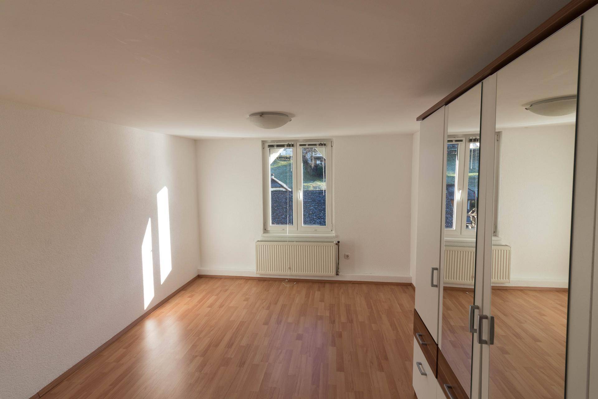 Helles Schlafzimmer mit Laminatboden, großem Fenster und einem Spiegelschrank, durchflutet von Sonnenlicht.