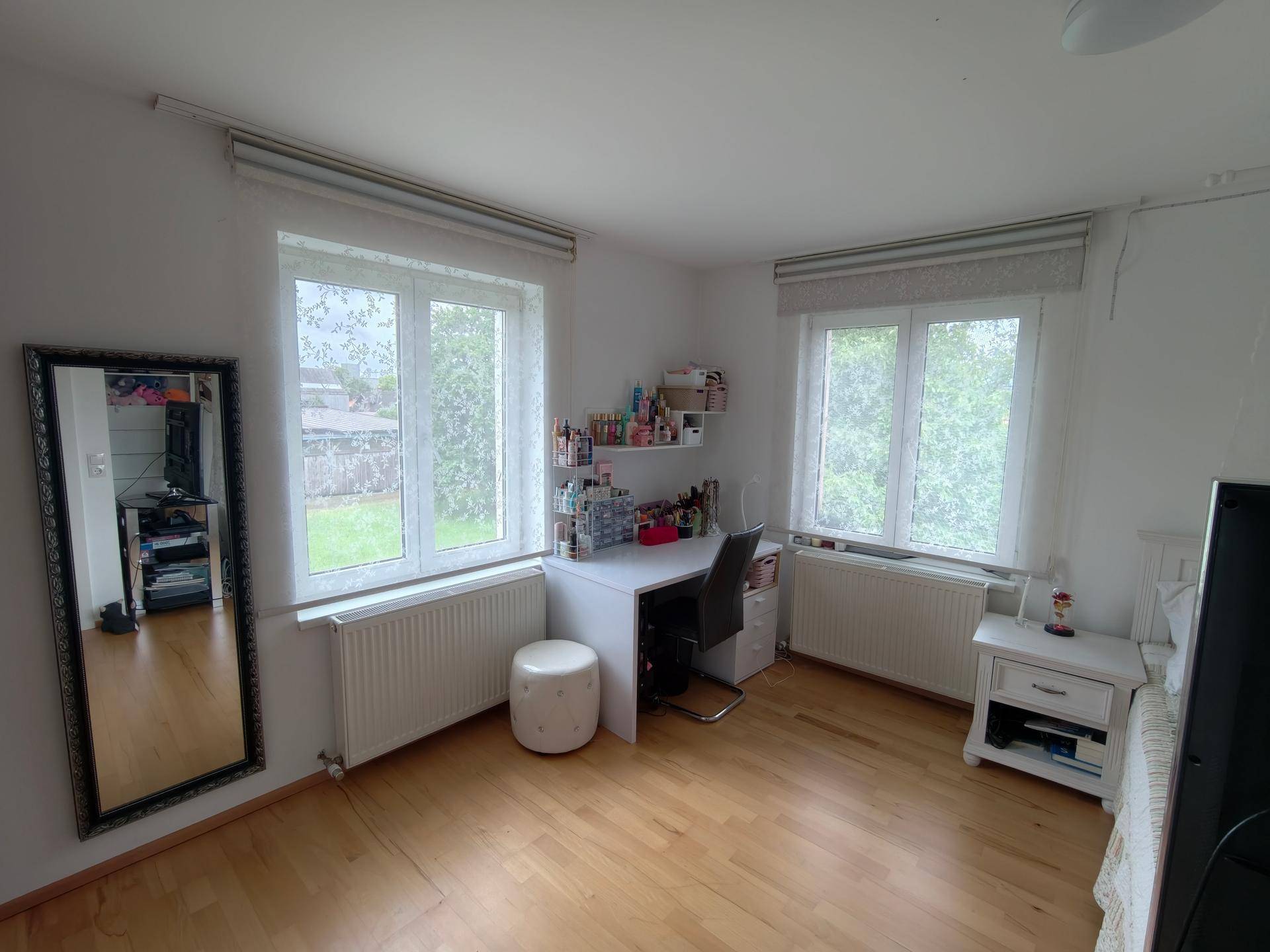 Helles Schlafzimmer mit Holzboden, Schreibtisch und zwei Fenstern mit Blick ins Grüne.