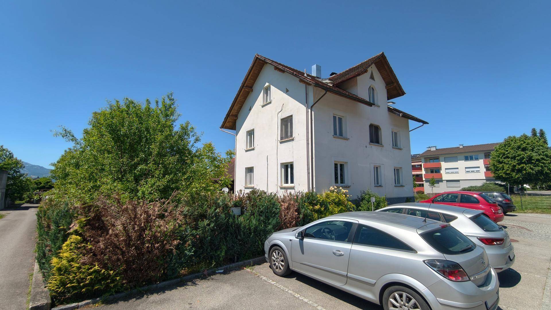 Ansicht des weißen Mehrfamilienhauses mit parkenden Autos und grüner Hecke an einem sonnigen Tag.