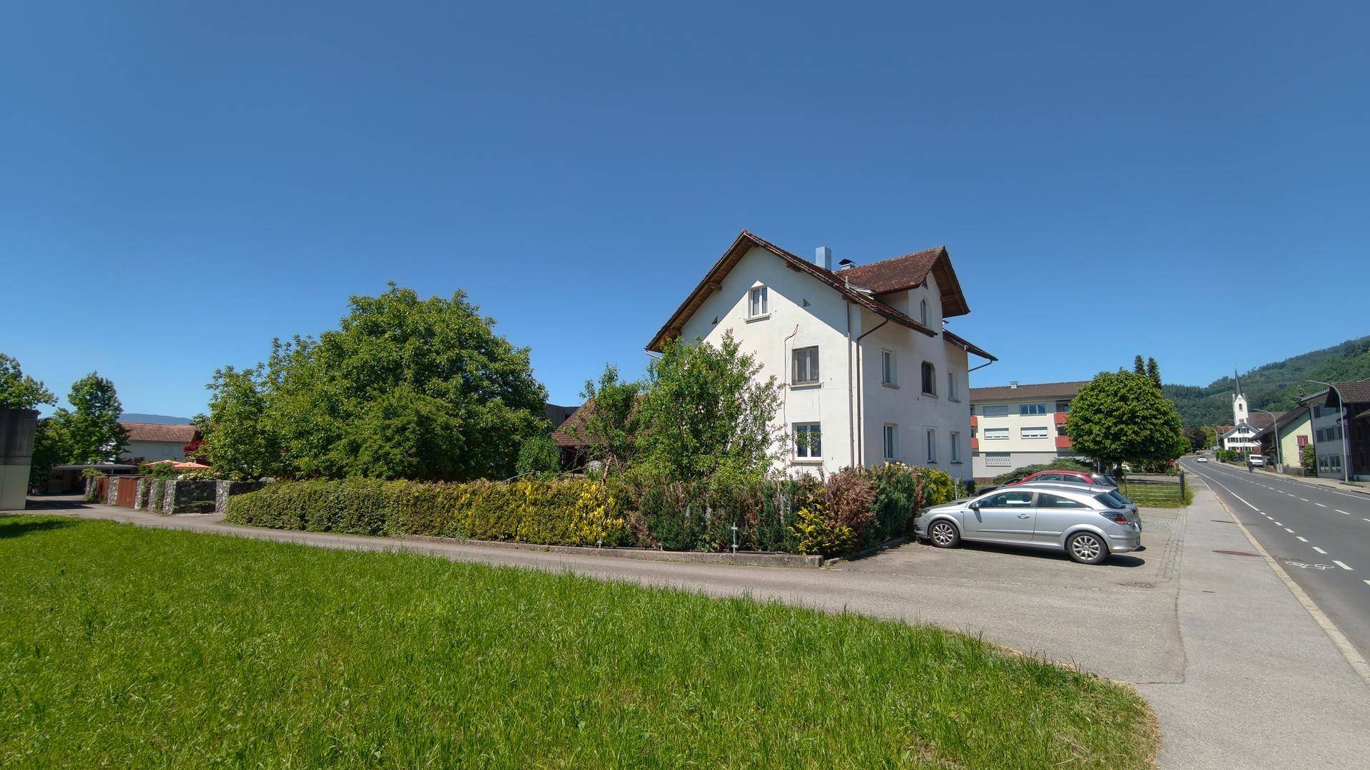 Mehrfamilienhaus an einer Straße mit Grünflächen und Bäumen unter strahlend blauem Himmel.