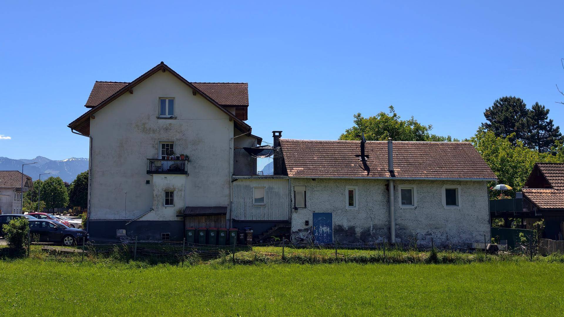 Seitenansicht des Mehrfamilienhauses mit Anbau und weitem Blick auf grüne Wiesen und Berge.