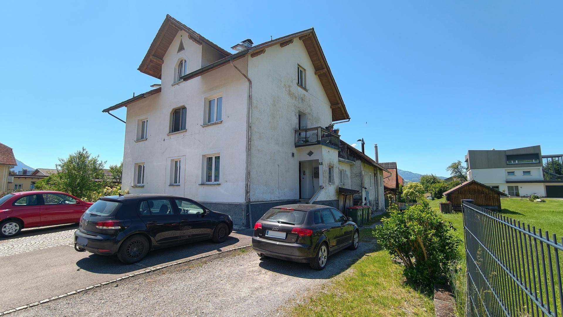 Rückansicht des Mehrfamilienhauses mit Balkon und geparkten Fahrzeugen auf dem Kiesparkplatz.