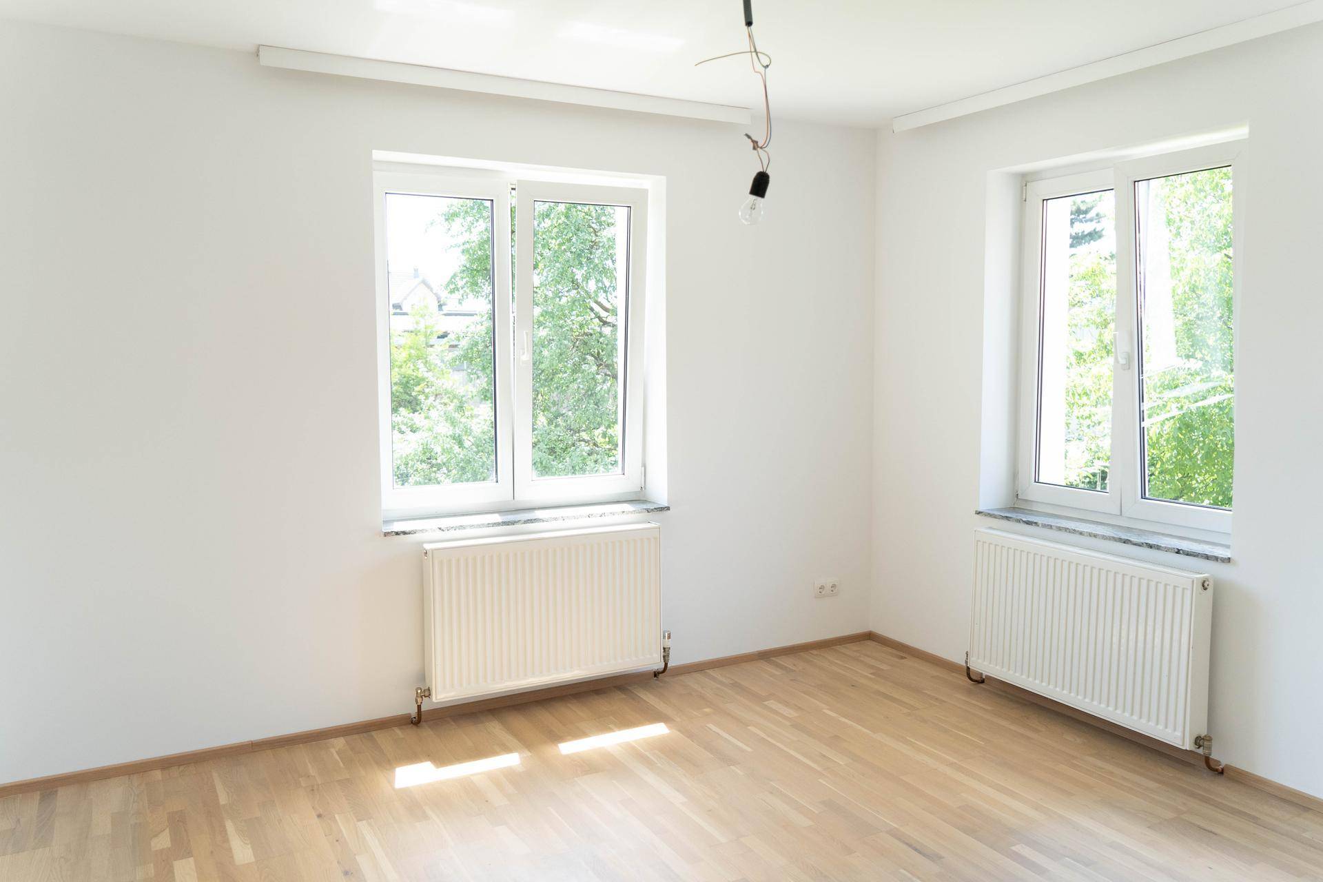 Helles Wohnzimmer mit zwei großen Fenstern, Heizkörpern und Holzboden.