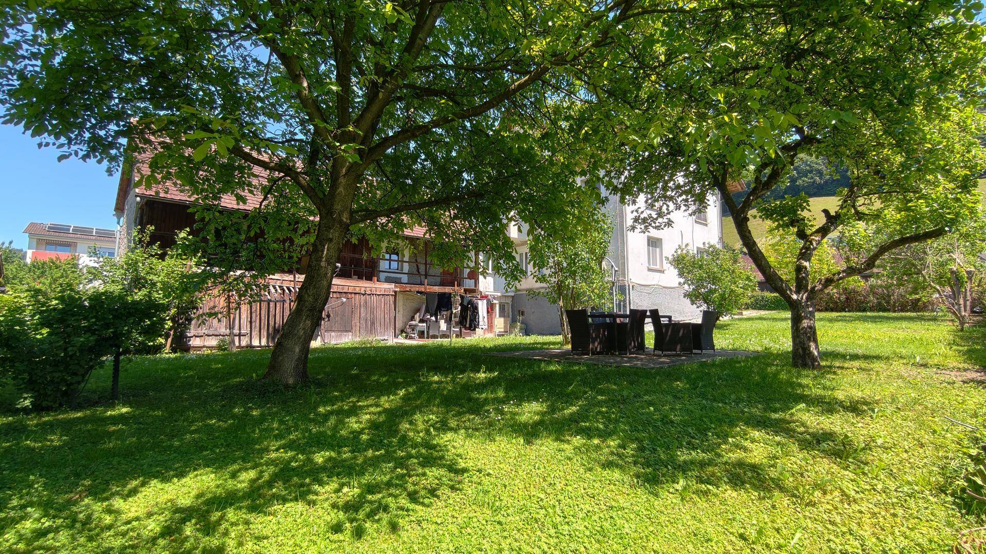 Weitläufiger Garten mit schattenspendenden Bäumen und einer möblierten Terrasse im Hintergrund.