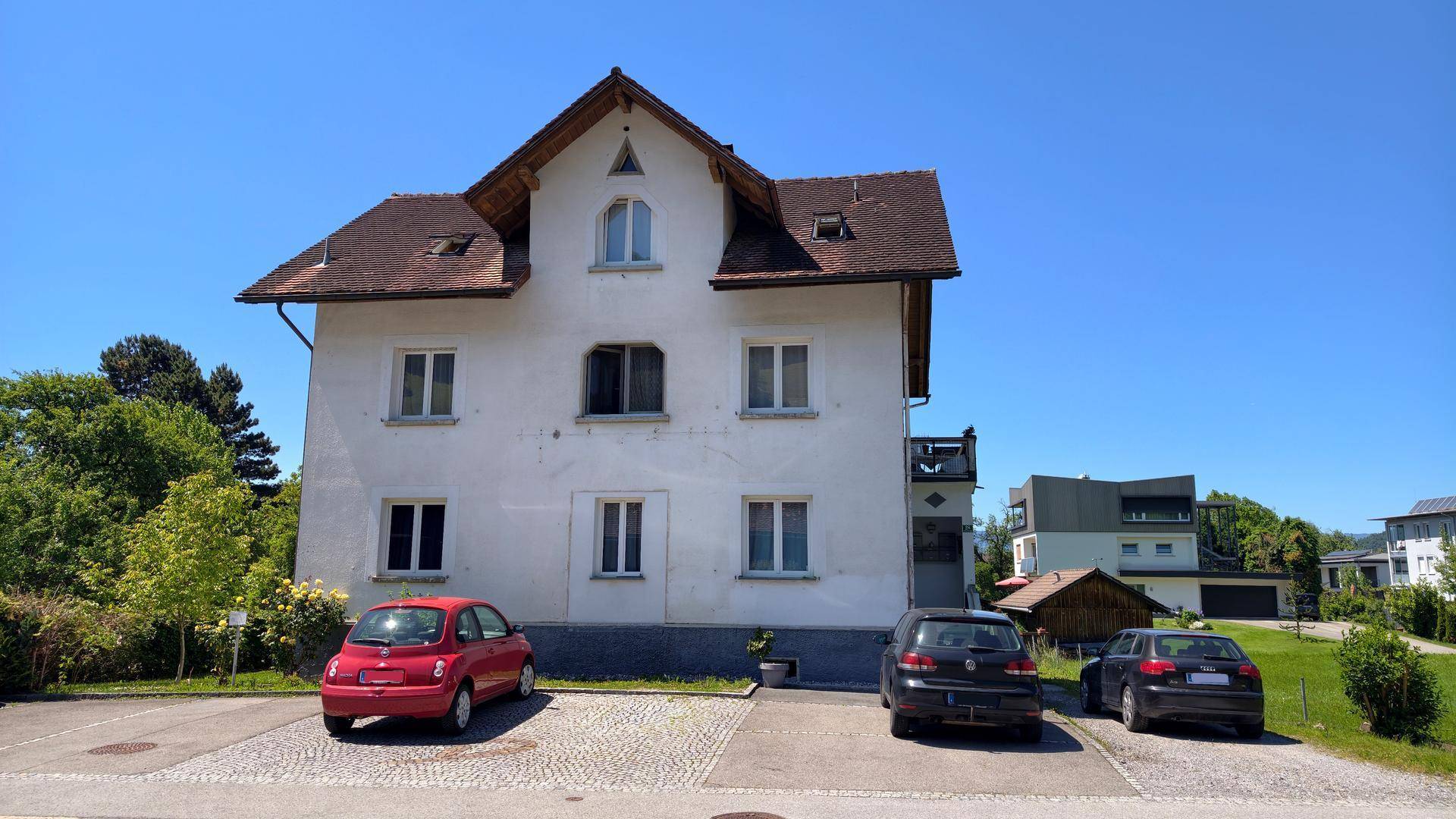 Vorderansicht des weißen Mehrfamilienhauses mit mehreren Fenstern und geparkten Autos auf dem Vorplatz.