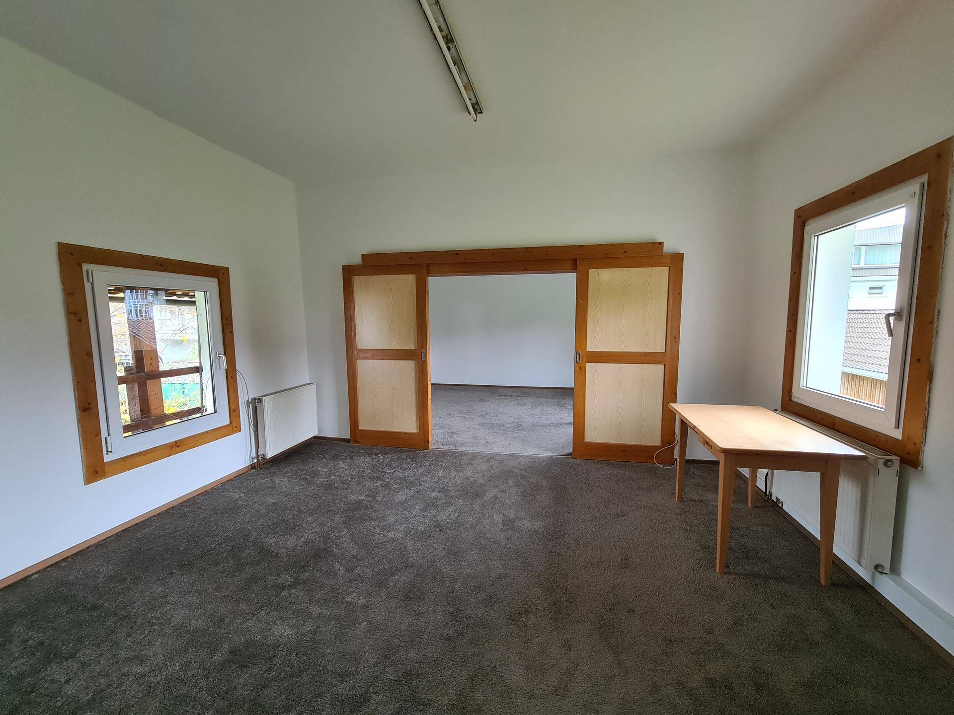 Geräumiges Zimmer mit grauem Teppichboden, Holzfenstern und großen Holzschiebetüren zu einem weiteren Raum.