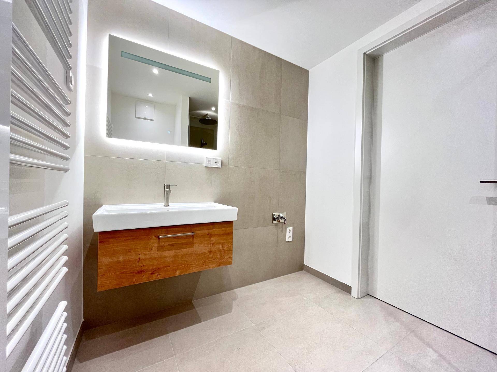 Modernes Badezimmer mit schwebendem Holzwaschtisch, beleuchtetem Spiegel und grauen Fliesen.