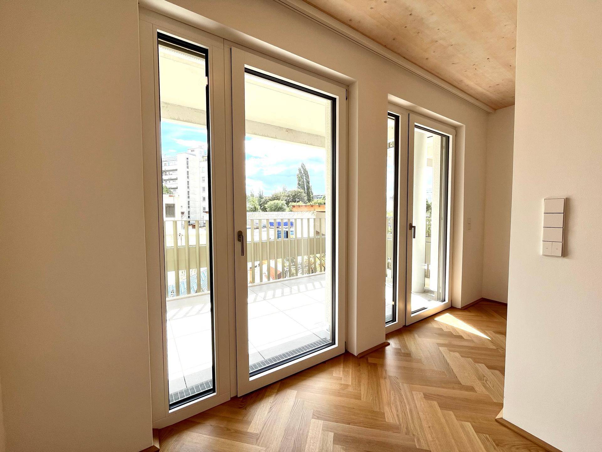 Heller Raum mit Fischgrätparkett, Holzbalkendecke und großen Fenstern zum Balkon.