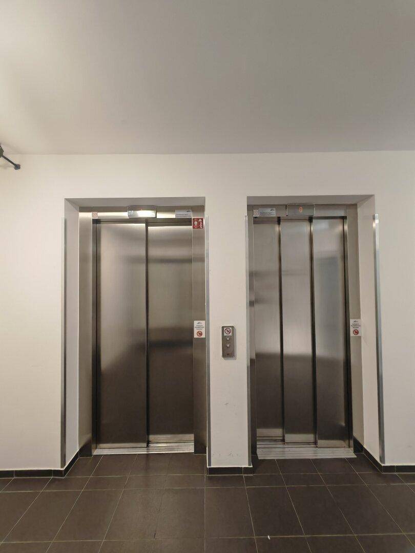 Moderner Flur mit zwei Aufzügen aus Edelstahl und dunklen Bodenfliesen.