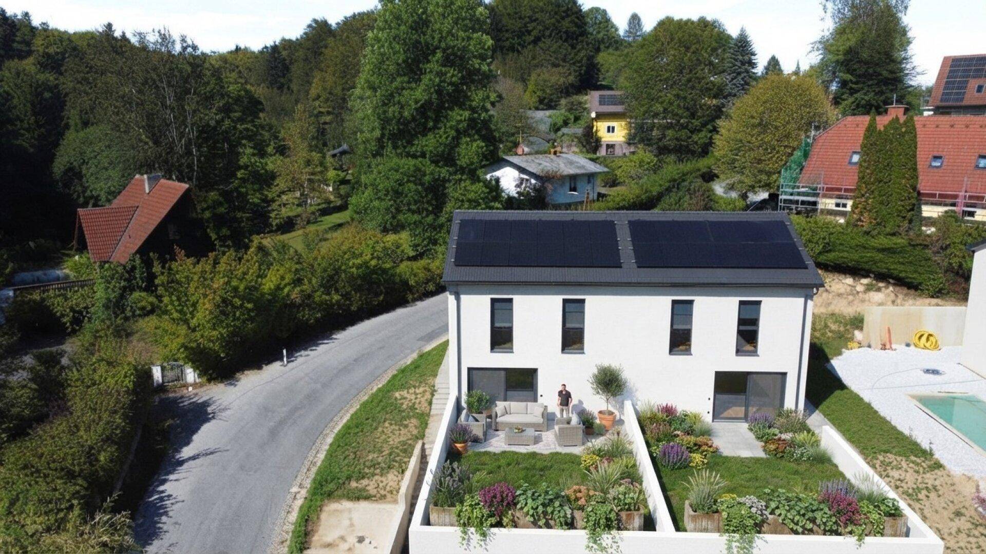 Modernes Haus mit Photovoltaikanlage, mehreren Terrassen und bepflanzten Hochbeeten in grüner Umgebung.