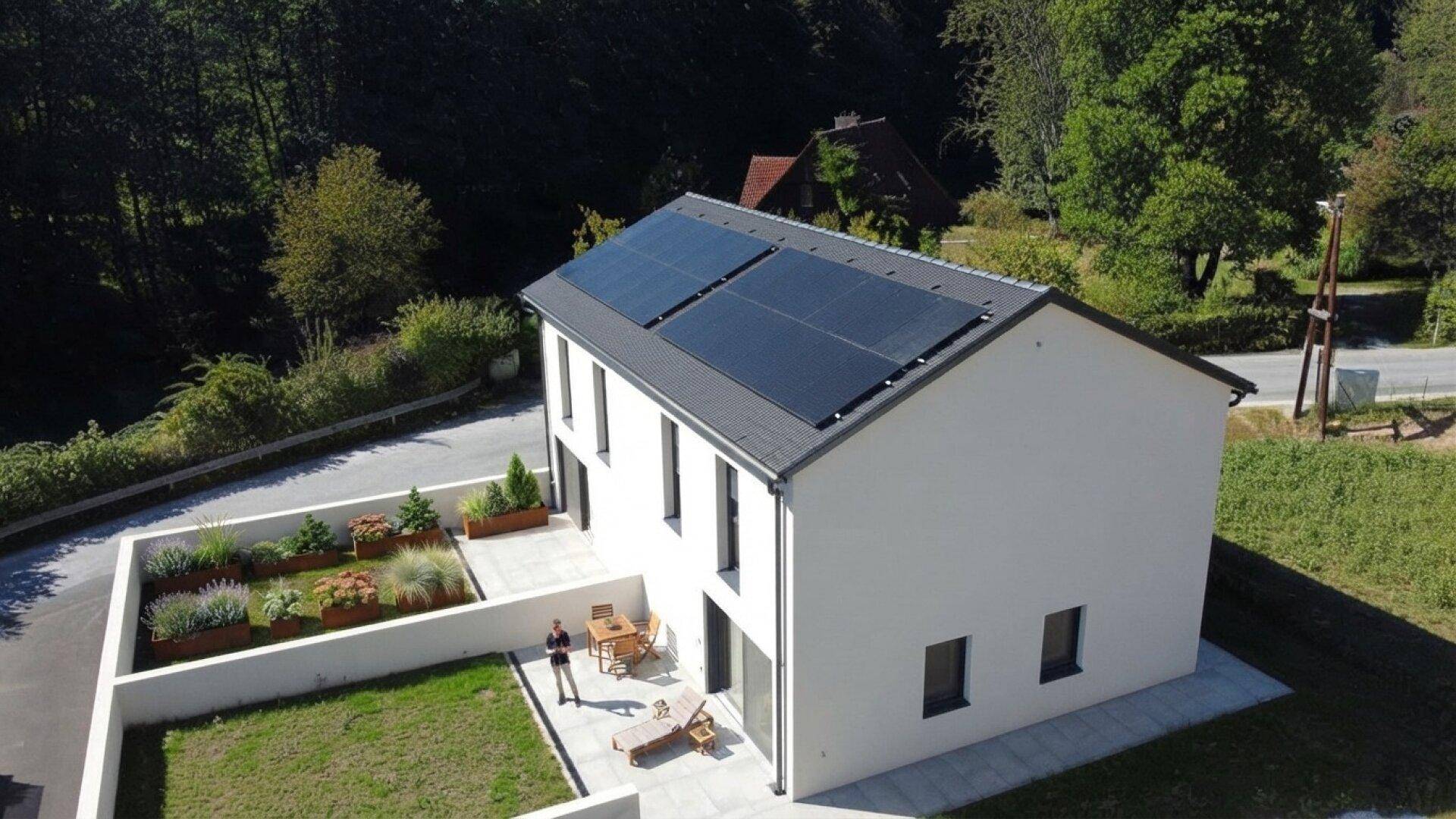 Modernes Einfamilienhaus mit Photovoltaikanlage, kleiner Terrasse und Rasenfläche in ländlicher Umgebung.