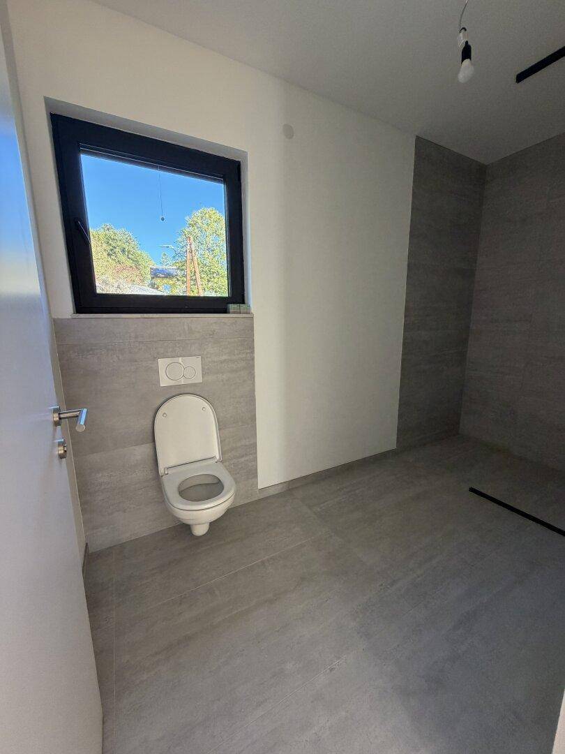 Modernes Badezimmer mit grauen Fliesen, wandhängendem WC und einem Fenster für Tageslicht.