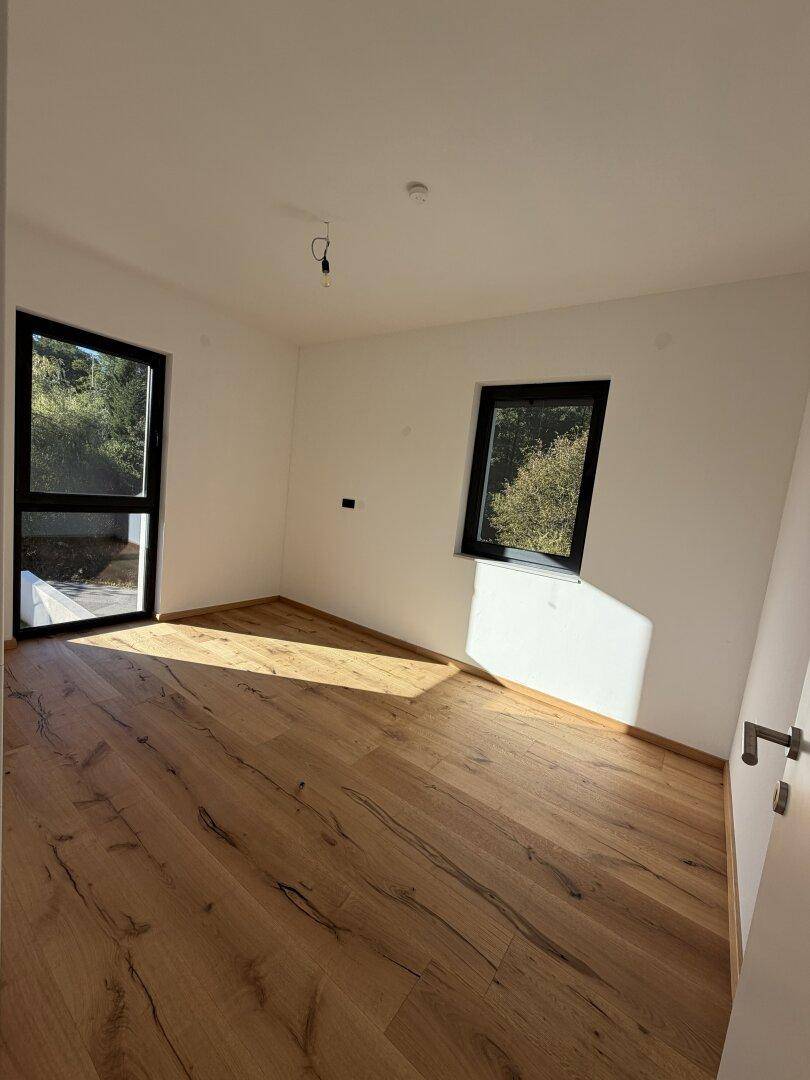 Lichtdurchfluteter Raum mit Holzdielenboden und großen Fenstern, die einen grünen Ausblick bieten.