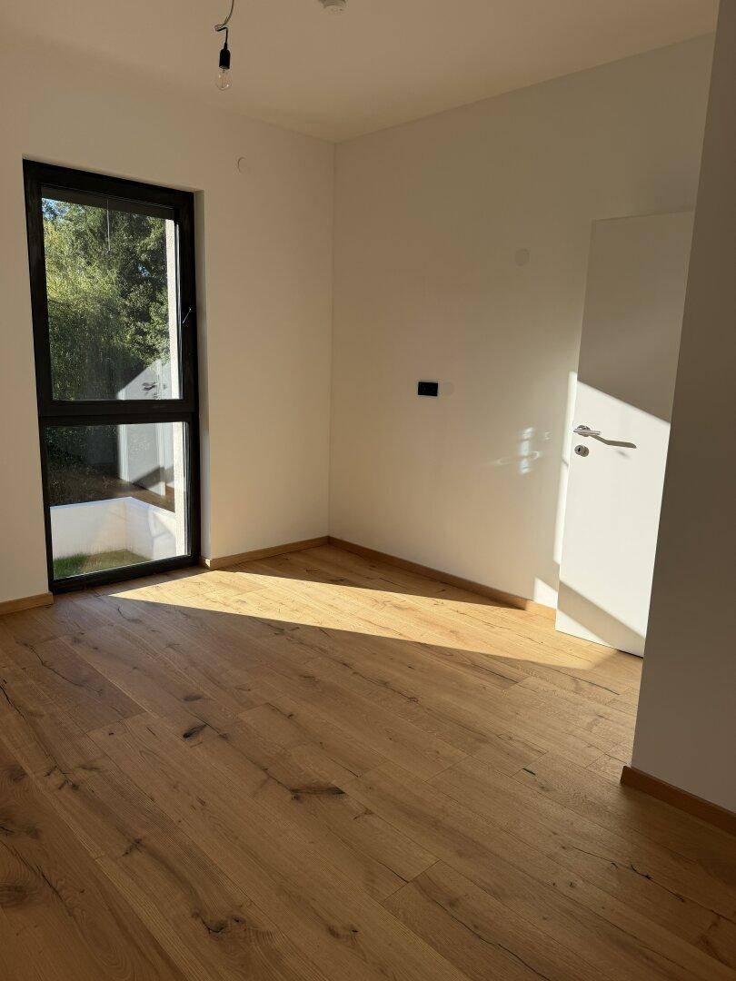 Sonniger Raum mit Holzdielenboden und großem Fenster, das viel natürliches Licht hereinlässt.