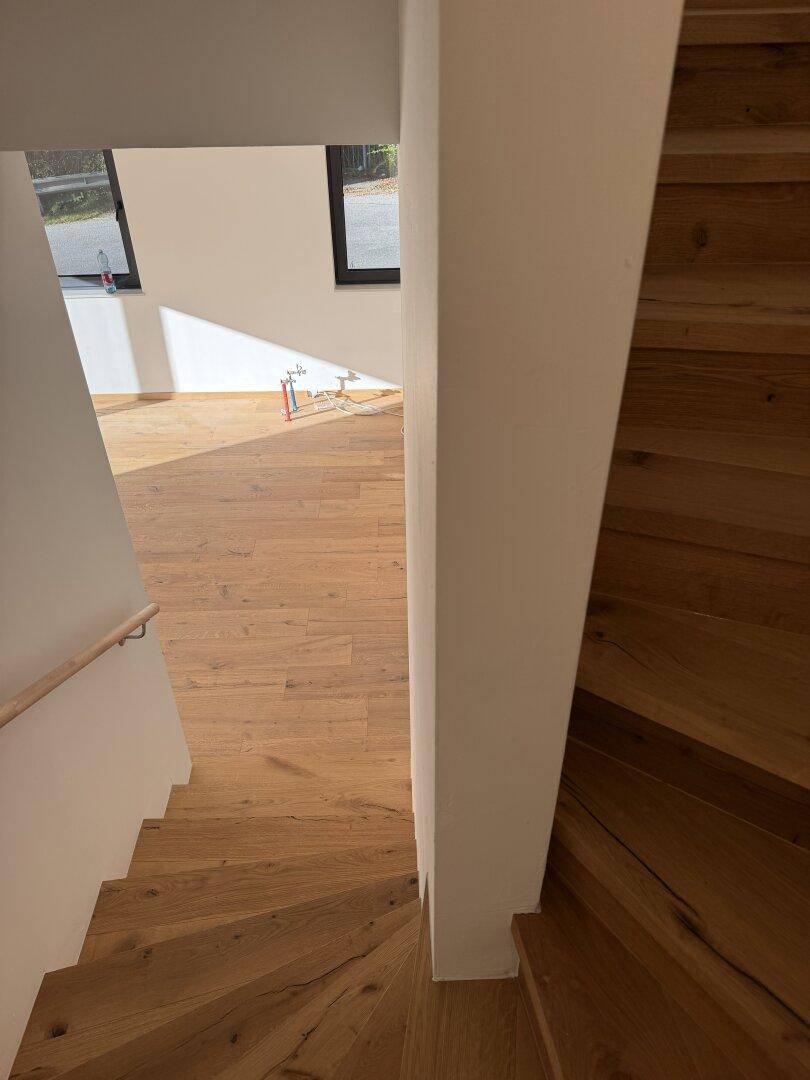 Blick von der Holztreppe hinunter in einen hellen Raum mit Holzdielenboden und Fenstern.