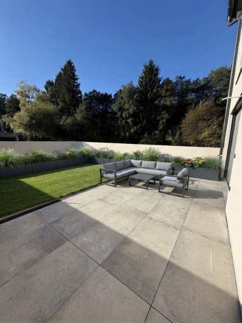 Moderne Terrasse mit großformatigen Fliesen, Rasenfläche und bequemen Outdoor-Sitzmöbeln.