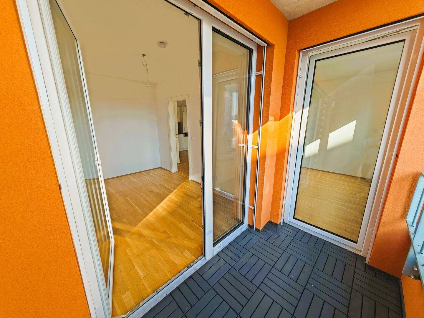 Balkon mit orangefarbenen Wänden und grauen Fliesen, zugänglich durch Glasschiebetüren.