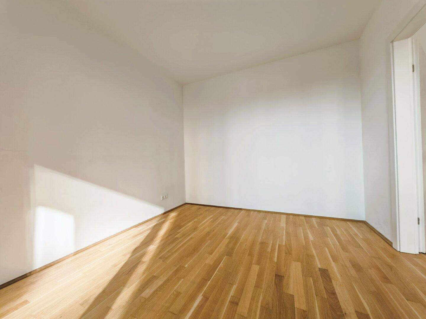 Leeres Zimmer mit hellem Parkettboden und weißen Wänden, gut beleuchtet durch Sonnenlicht.