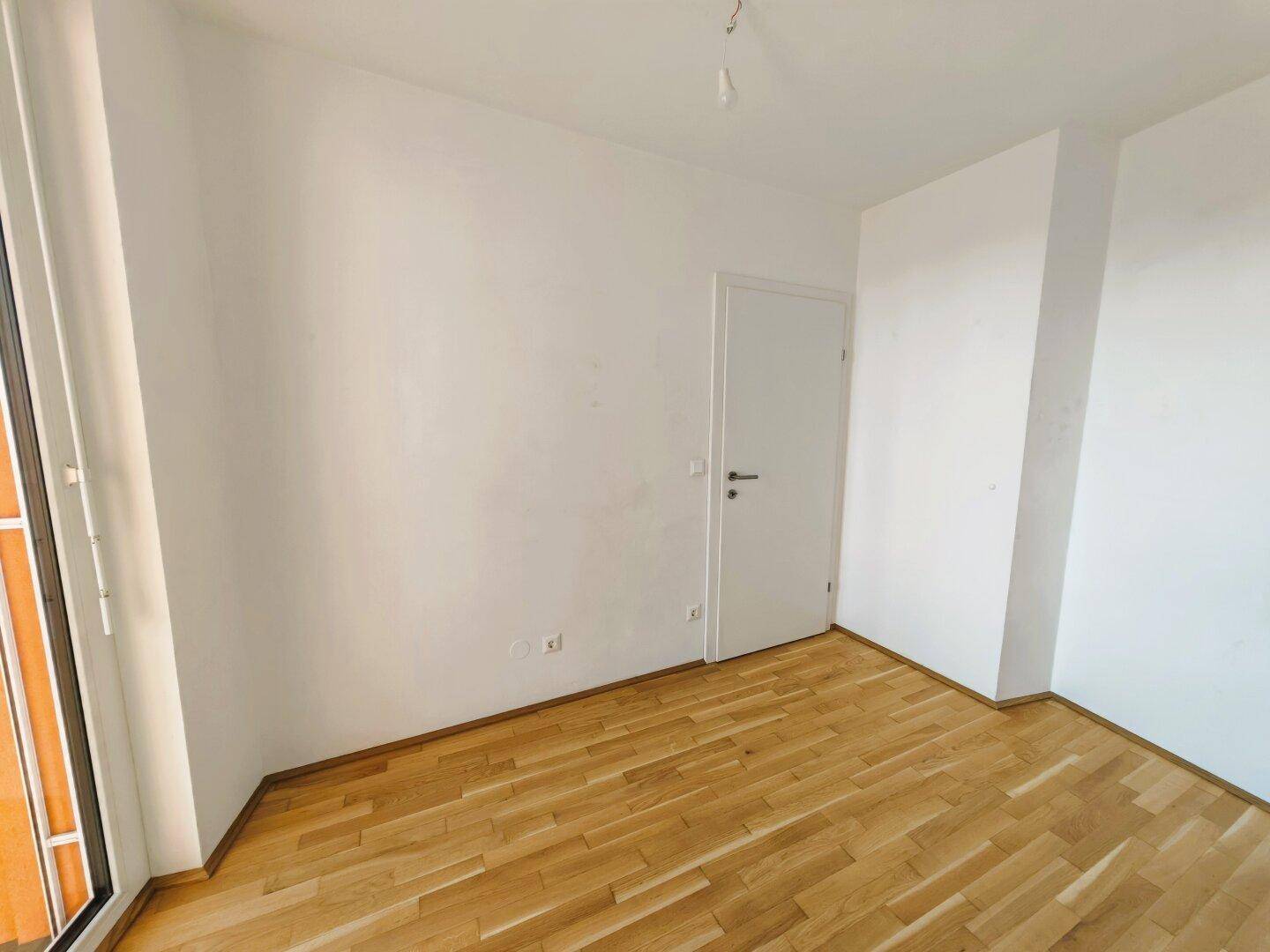Leerer Raum mit Parkettboden und weißer Tür, der auf weitere Bereiche führt.