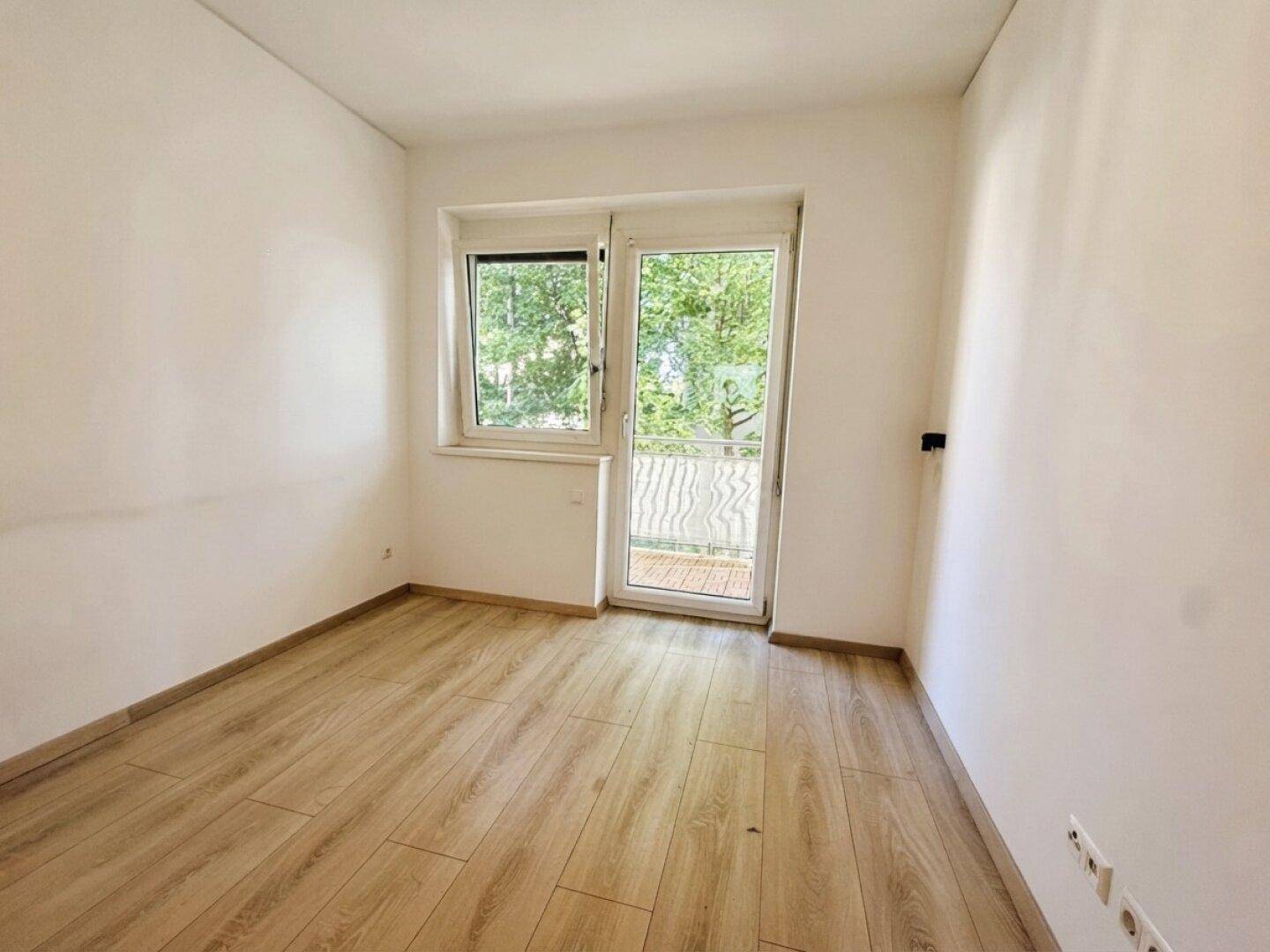 Leeres Zimmer mit Holzboden und großem Fenster, das viel Tageslicht hereinlässt.