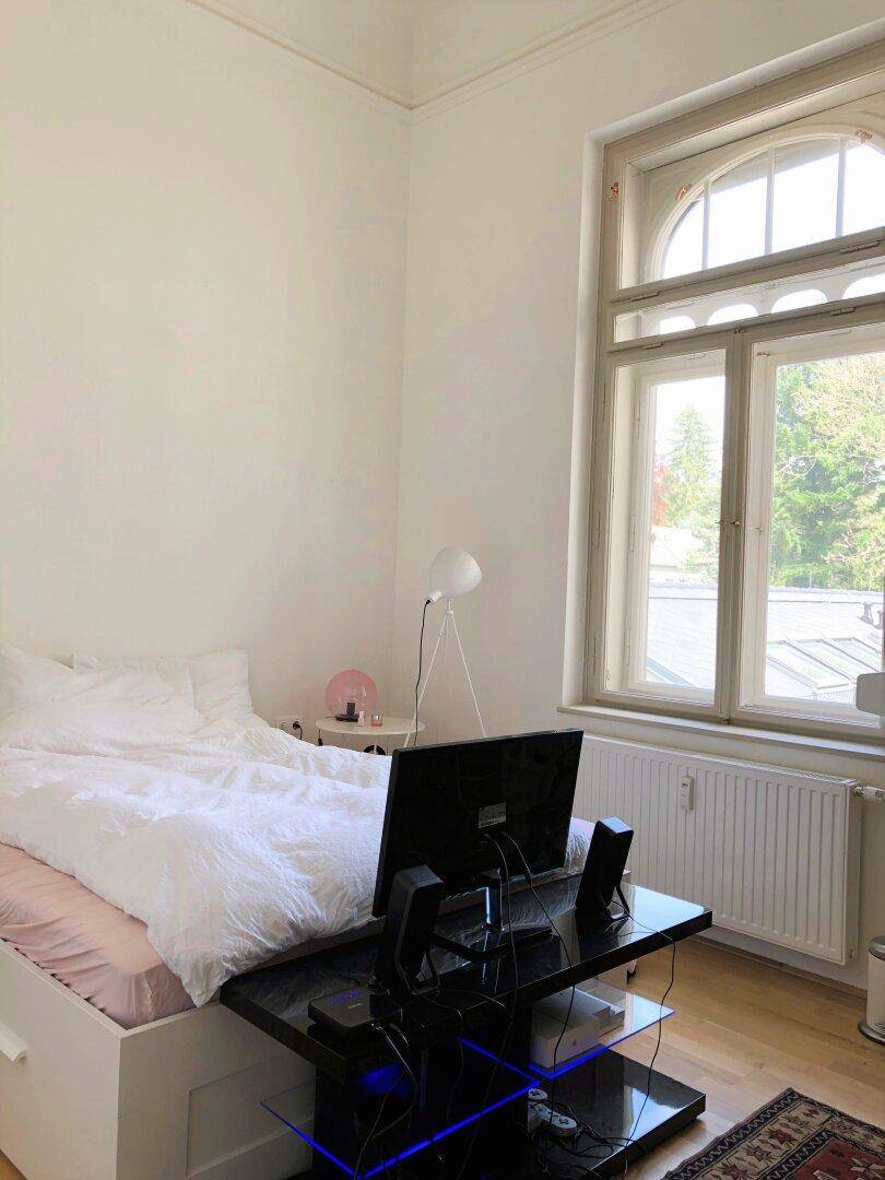 Schlafzimmer mit Holzboden, hohen Decken und einem großen Fenster mit Ausblick ins Grüne.