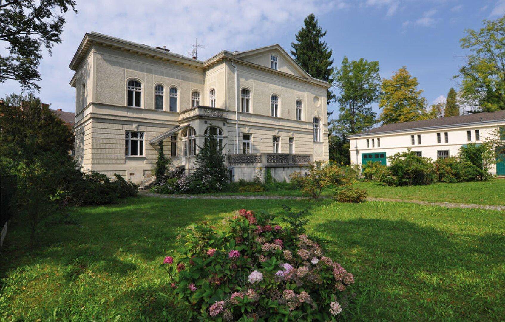 Historische Villa mit weitläufigem Garten und altem Baumbestand in grüner Umgebung.