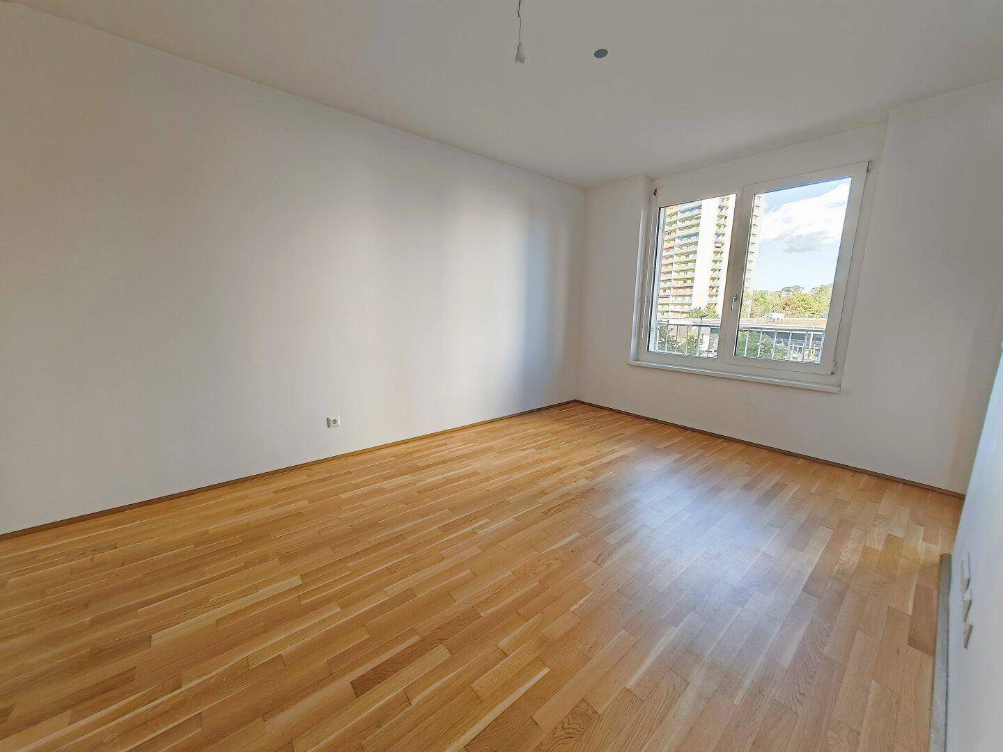 Helles Zimmer mit Parkettboden und großem Fenster, das viel Tageslicht hereinlässt.