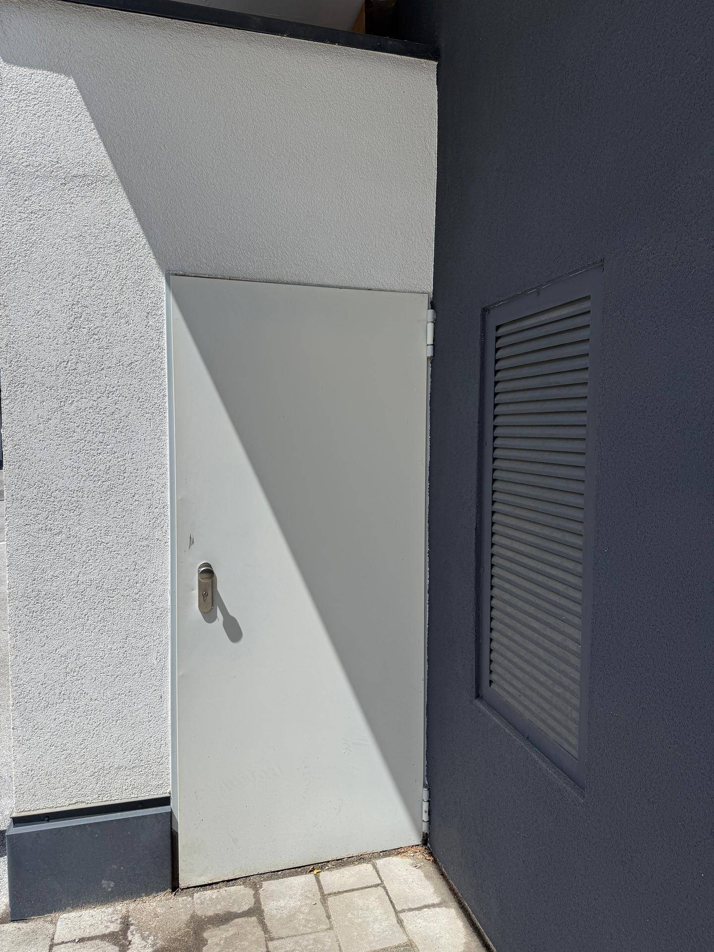 Weiße Außentür neben einer dunkelgrauen Wand mit Lüftungsgitter.