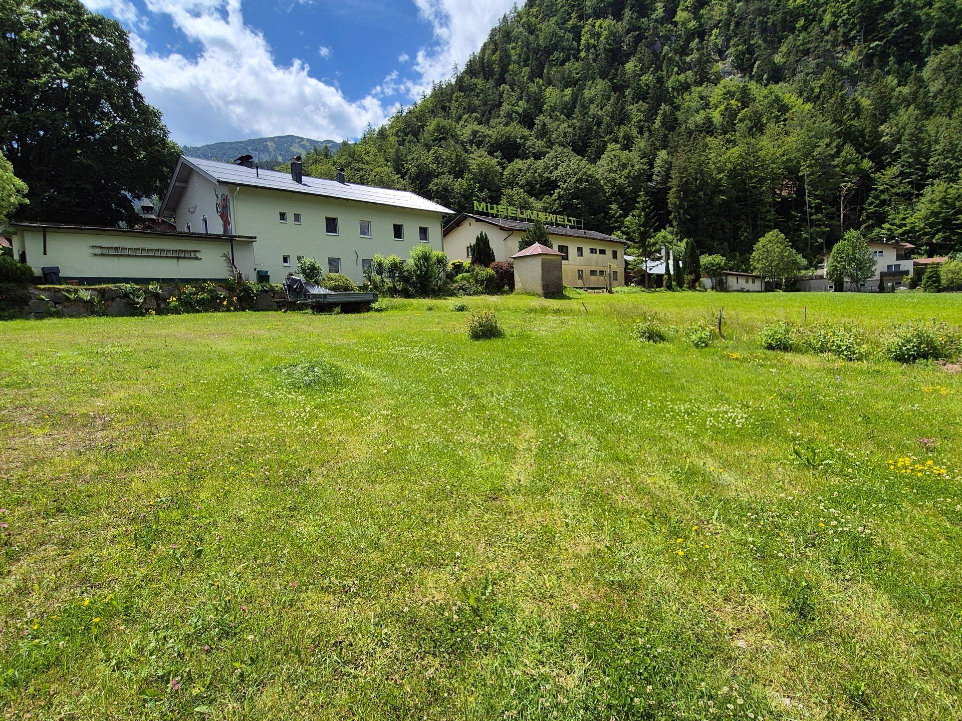 Große Wiesenfläche vor einem Gebäude mit Solaranlage und bewaldeten Bergen im Hintergrund.