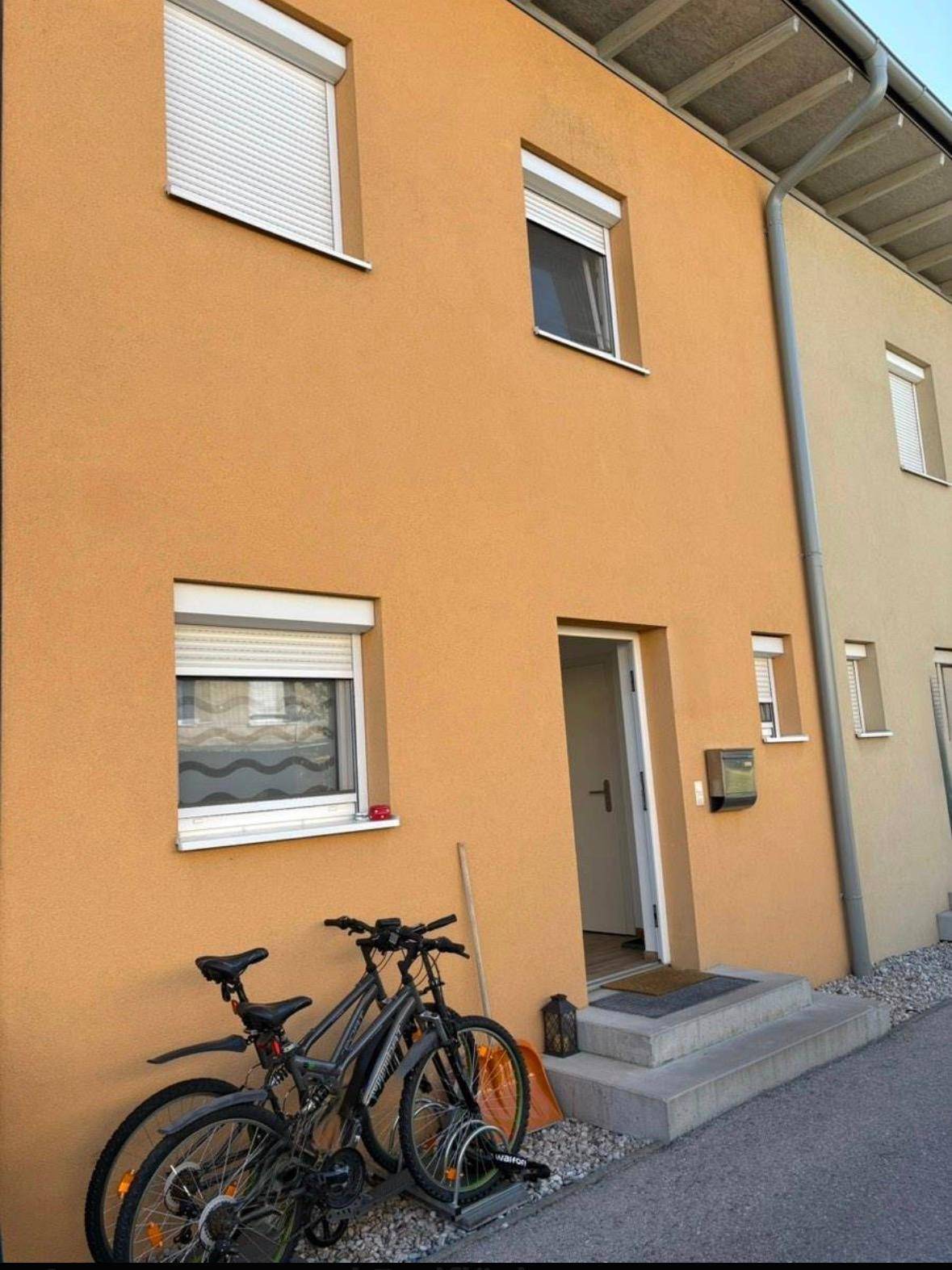 Vorderansicht eines Reihenhauses mit orangefarbener Fassade, weißen Rollläden und Eingangsbereich.