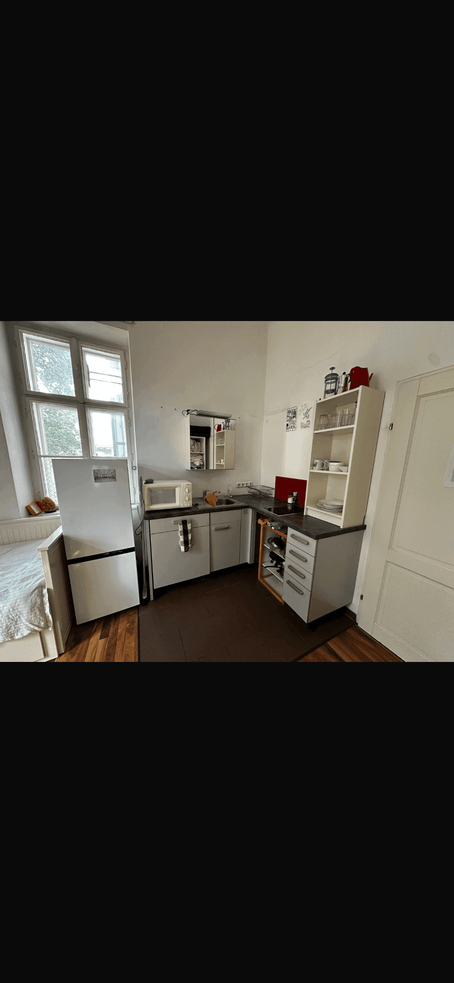 Funktionale Küchenzeile mit Kühlschrank, Mikrowelle und Spüle, gut beleuchtet durch ein großes Fenster.