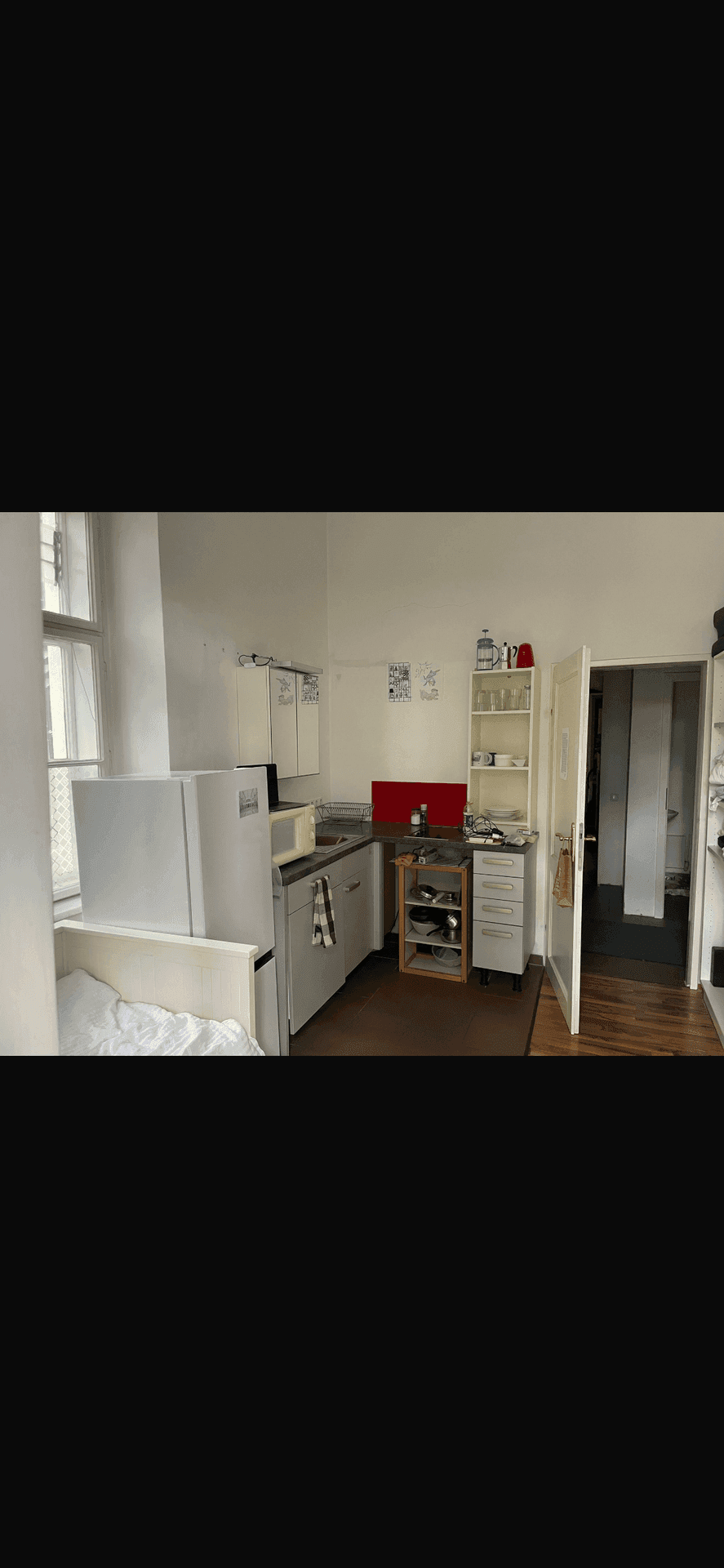 Kleine Küchenzeile mit Kühlschrank, Spüle und Regalen, integriert in einen Wohnraum mit Schlafbereich.