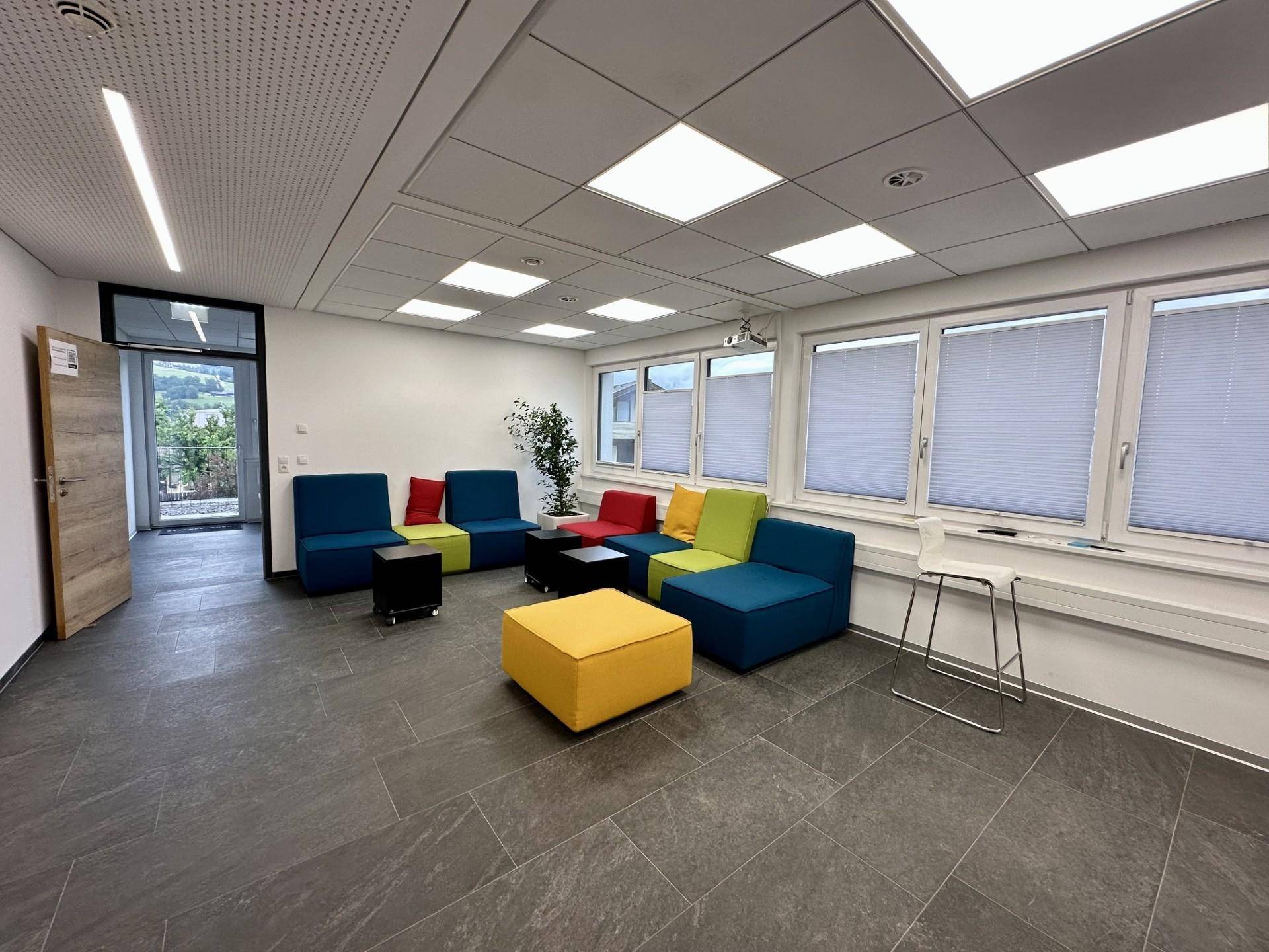 Großer Lounge-Bereich mit farbenfrohen Sitzmöbeln, vielen Fenstern und Zugang nach draußen.