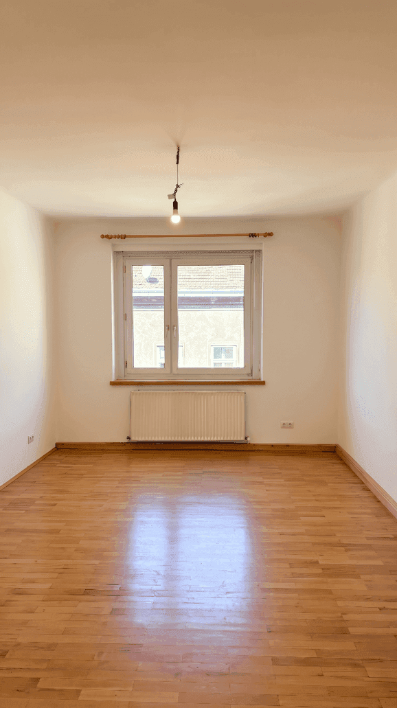 Heller, leerer Raum mit Parkettboden und einem Fenster, das den Raum mit natürlichem Licht durchflutet.