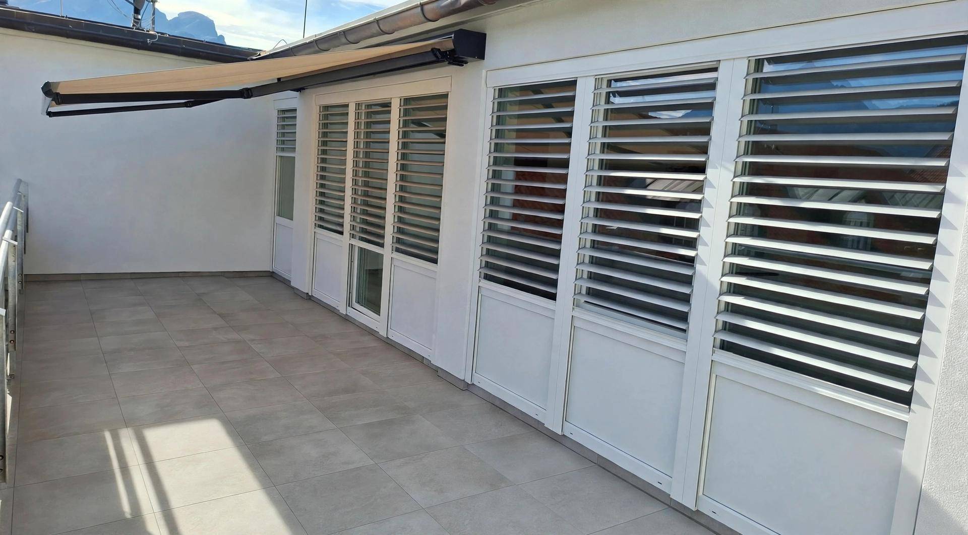 Geräumige Terrasse mit Fliesenboden, weißen Fensterläden und ausfahrbarer Markise für Sonnenschutz.