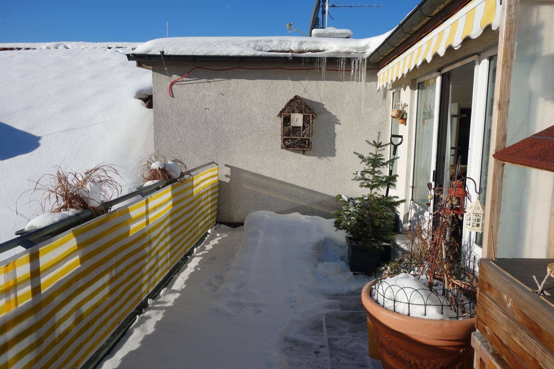 Balkon im Winter mit Schnee, gelb-weißem Sichtschutz und Topfpflanzen unter blauem Himmel.