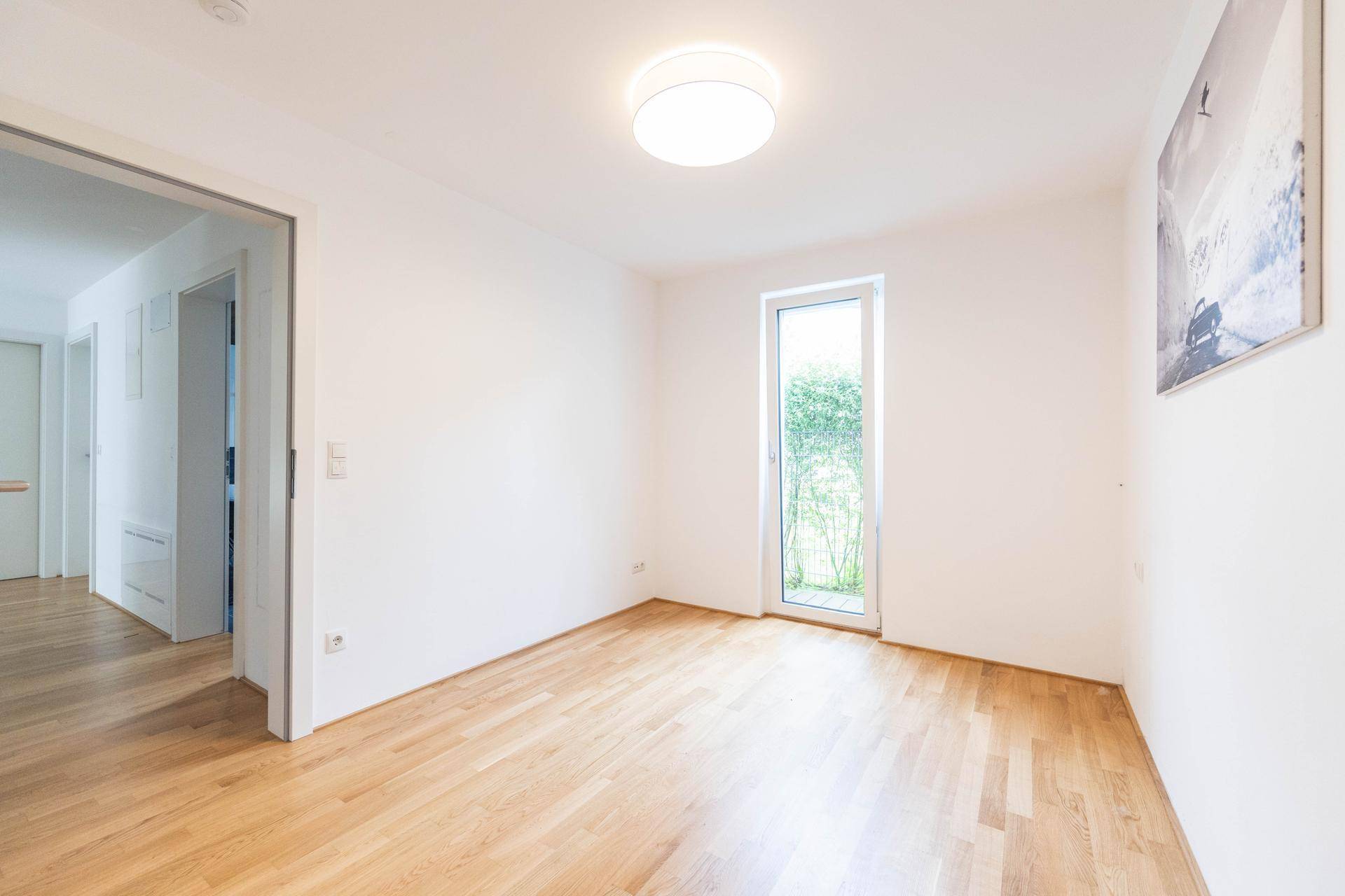 Lichtdurchflutetes Zimmer mit hellem Holzparkett und hoher Fenstertür, die nach draußen führt.