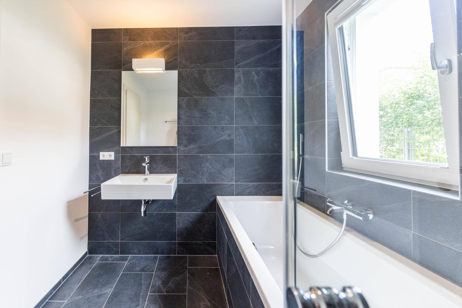Modernes Badezimmer mit dunklen Fliesen, Badewanne, Dusche und einem Fenster für Tageslicht.